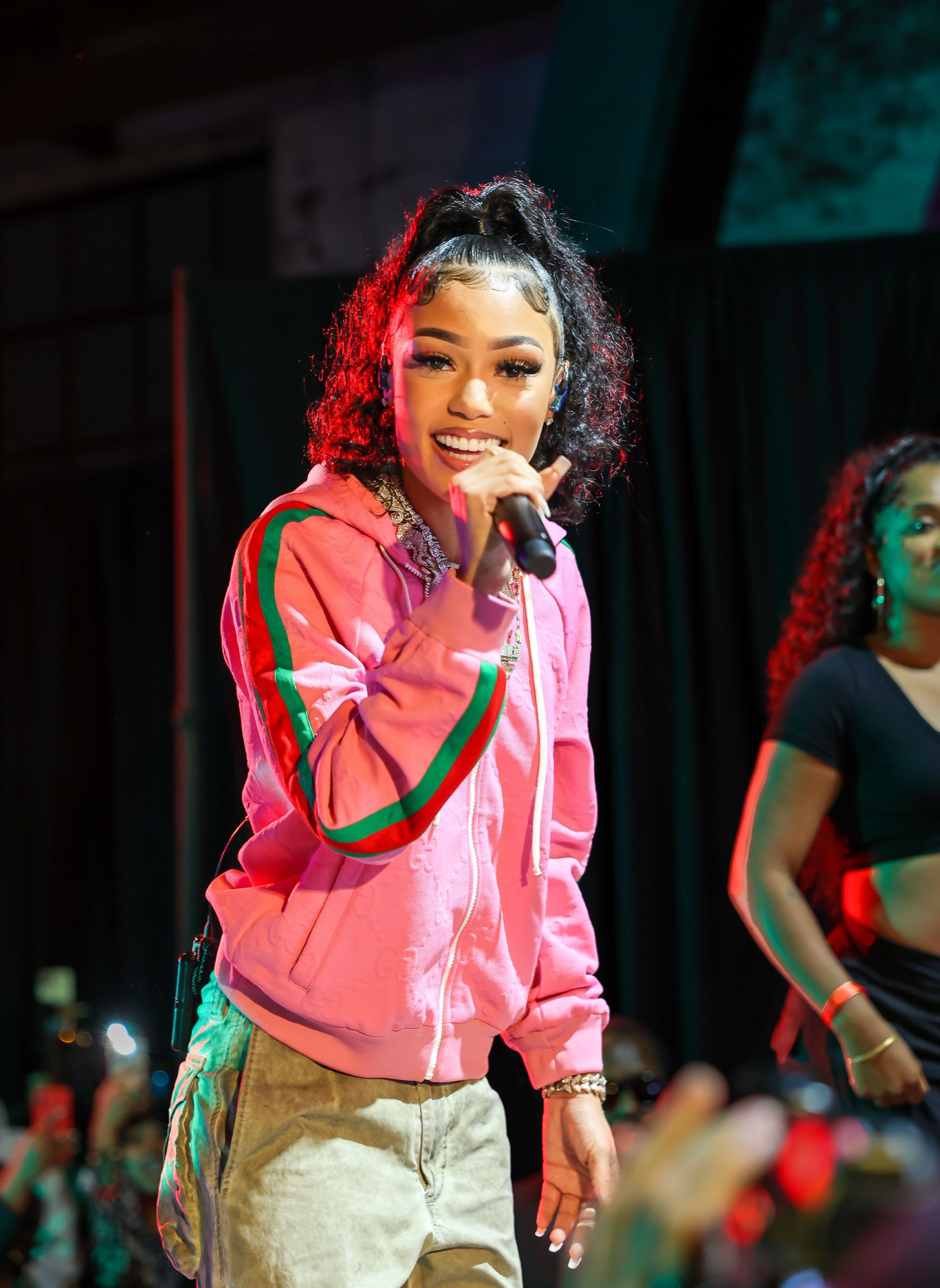Performer in pink hoodie singing on stage with colorful lighting effects.