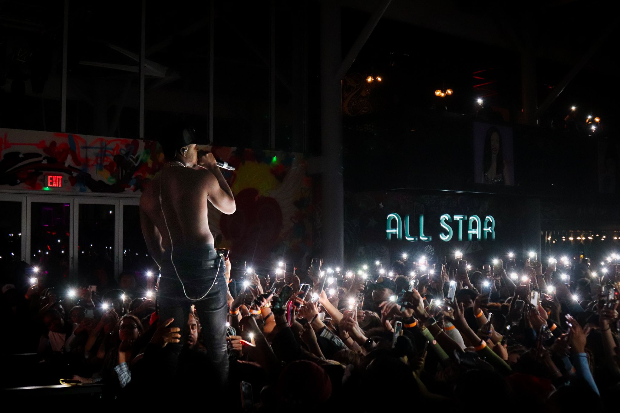 Performer on stage silhouetted against crowd holding up phone lights with 'All Star' sign in background.