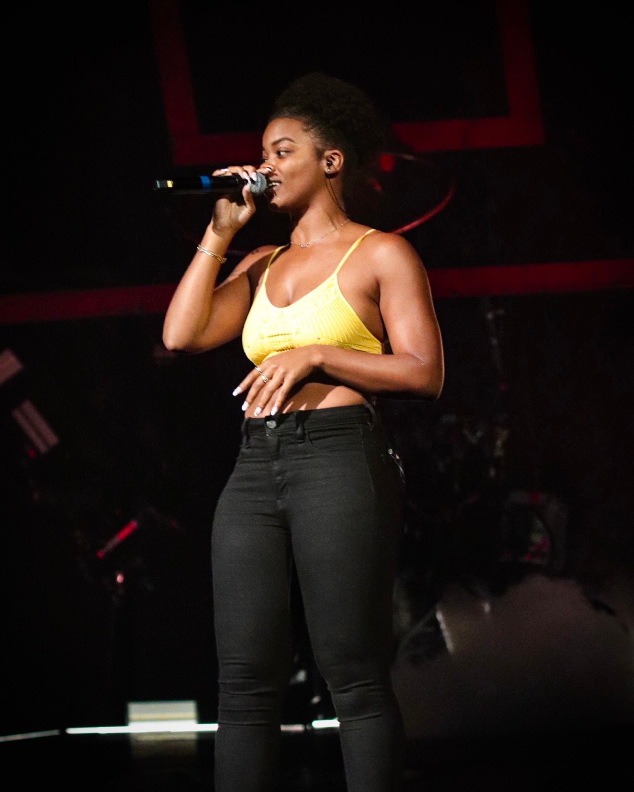 Singer in yellow tank top and black jeans performs on a dark stage with microphone.