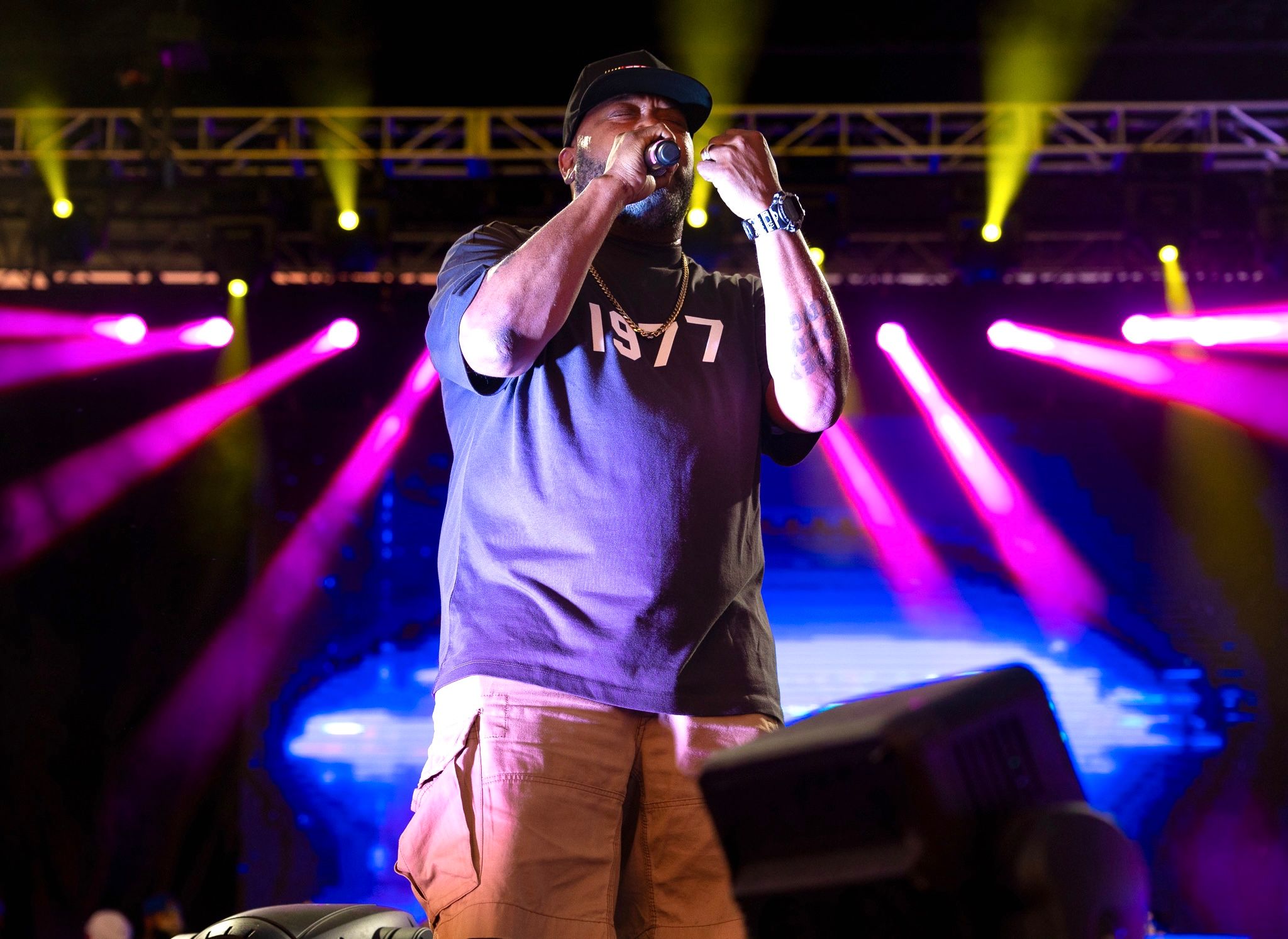 Concert performer in gray t-shirt singing on stage with dramatic purple and yellow lighting.