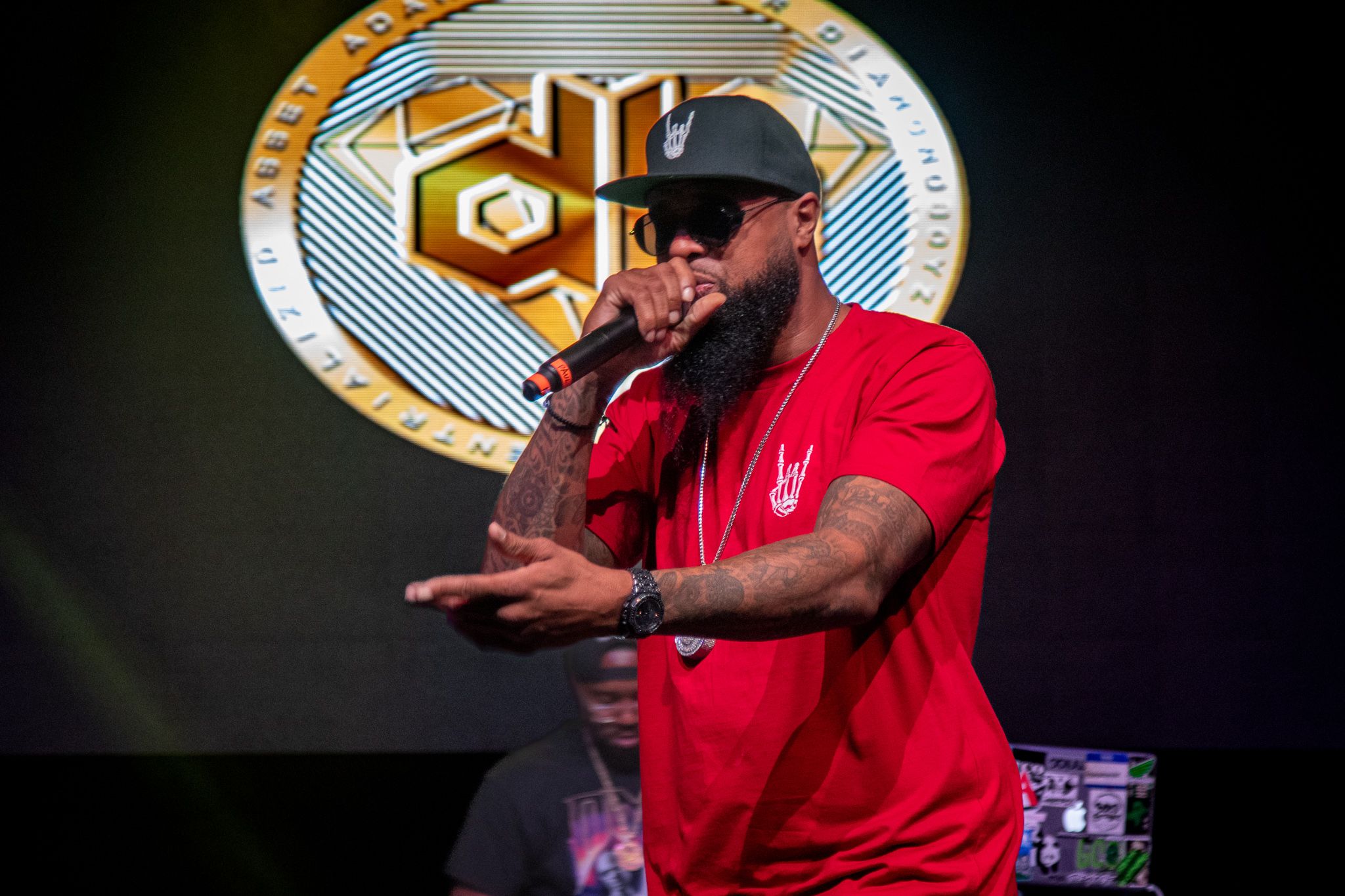 Performer in red shirt and cap gesturing while rapping in front of geometric logo projection.