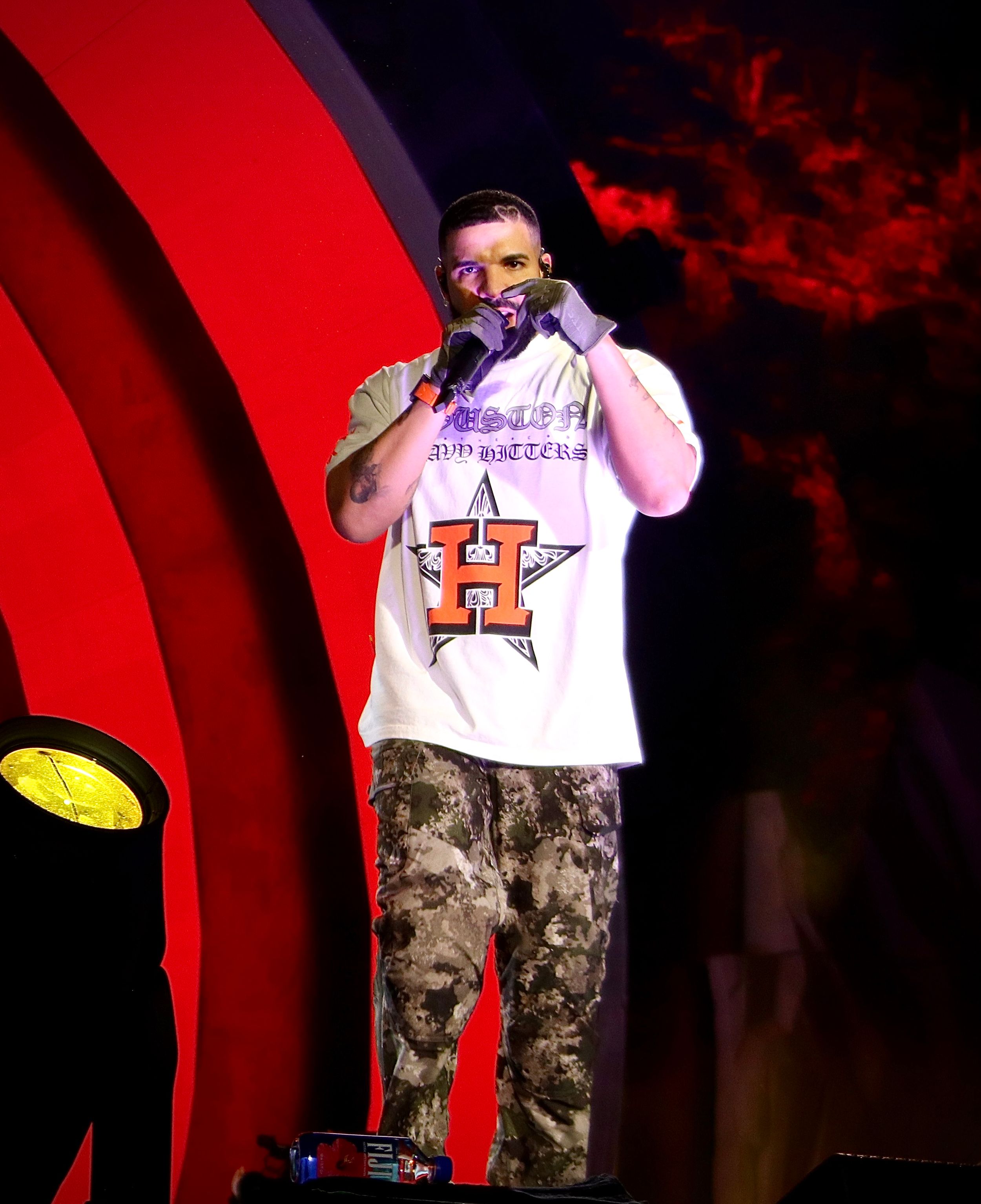 Performer on stage wearing white t-shirt and camo pants against red backdrop during concert.