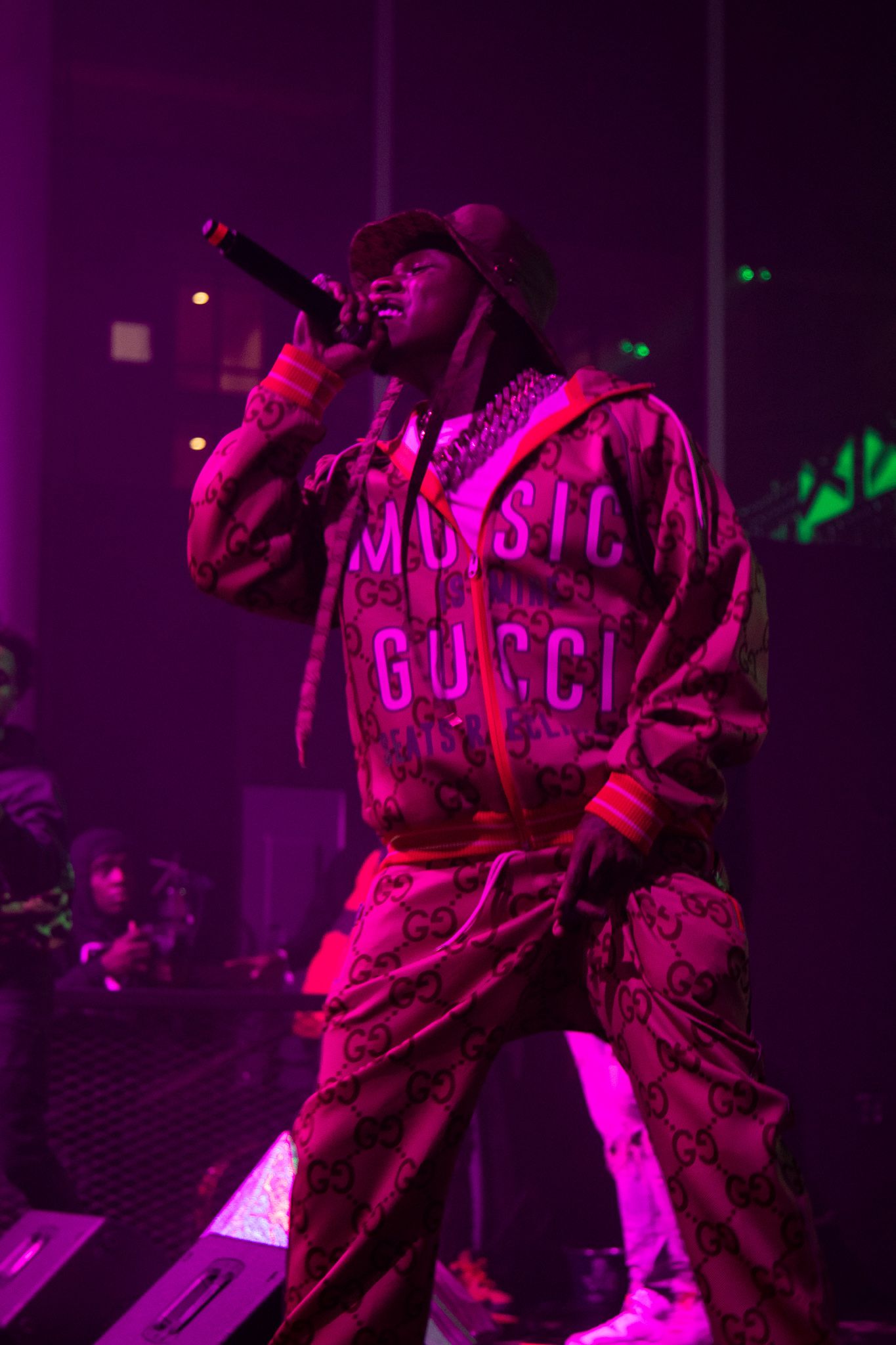 Concert performer in pink Gucci outfit on stage with purple lighting effects.