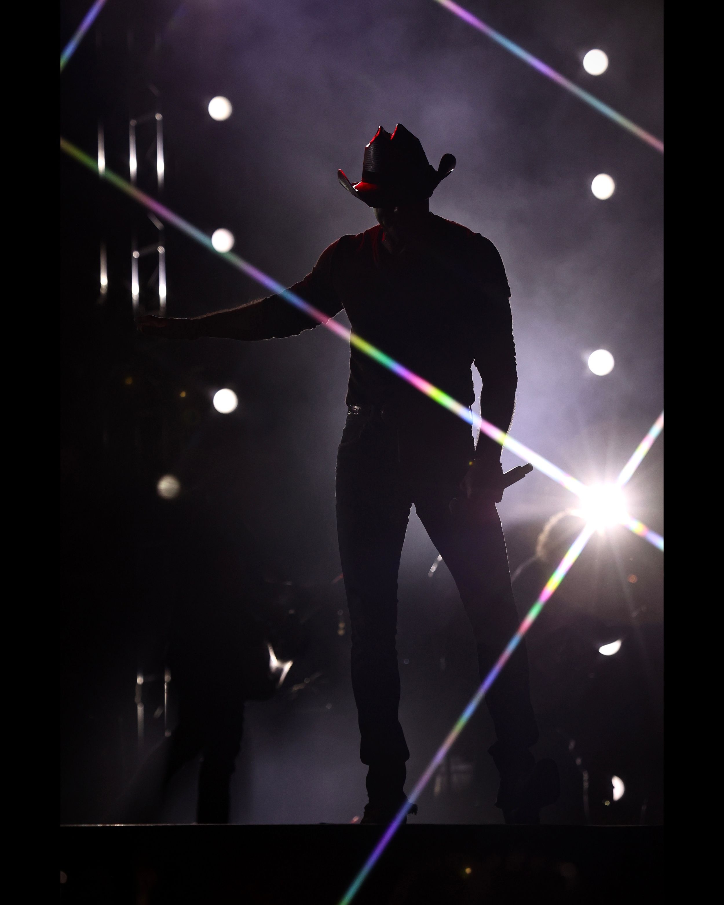 Dramatic silhouette of performer in cowboy hat against bright stage lights and beams.