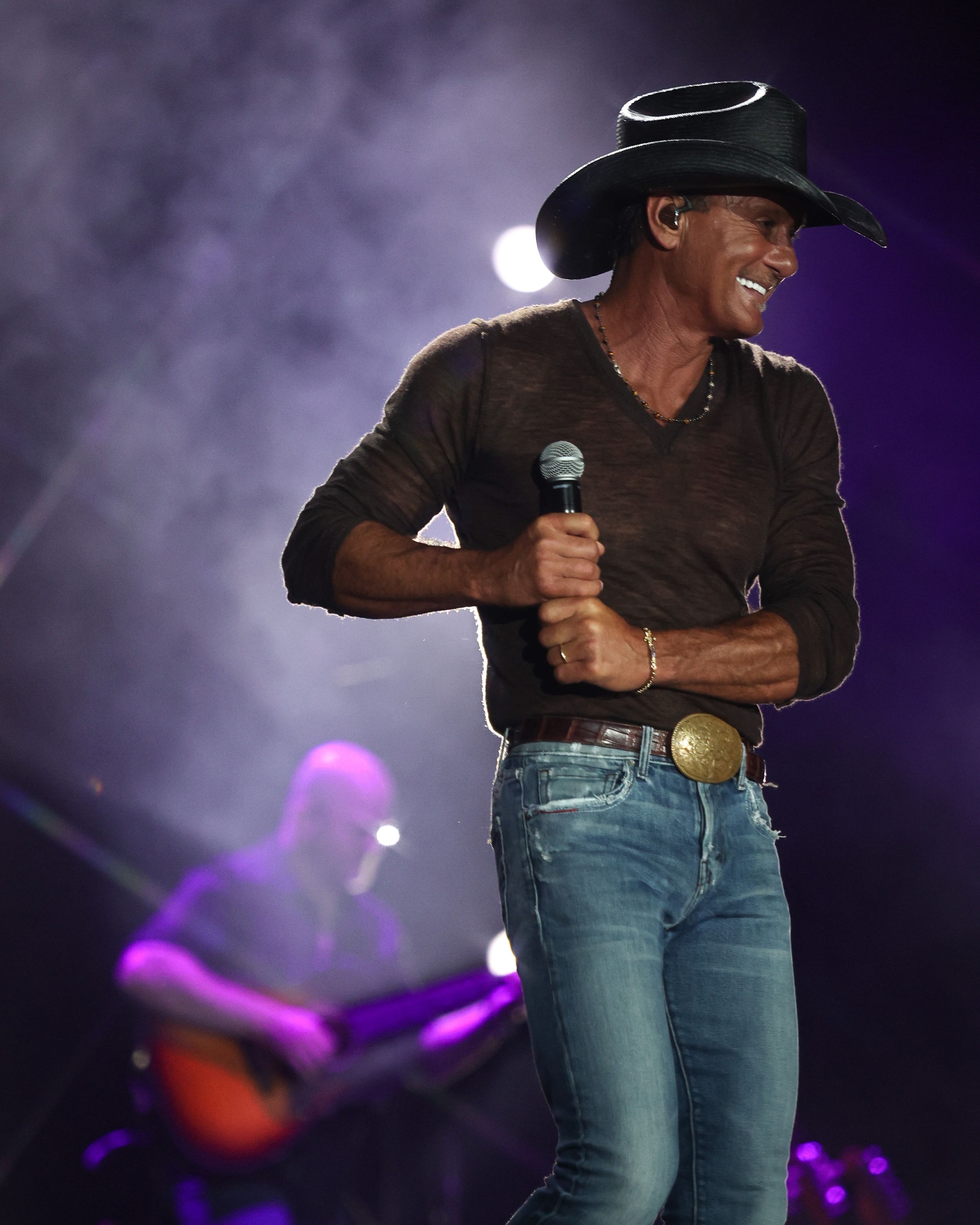 Country music performer in black cowboy hat and v-neck shirt sings on stage with purple lighting effects.