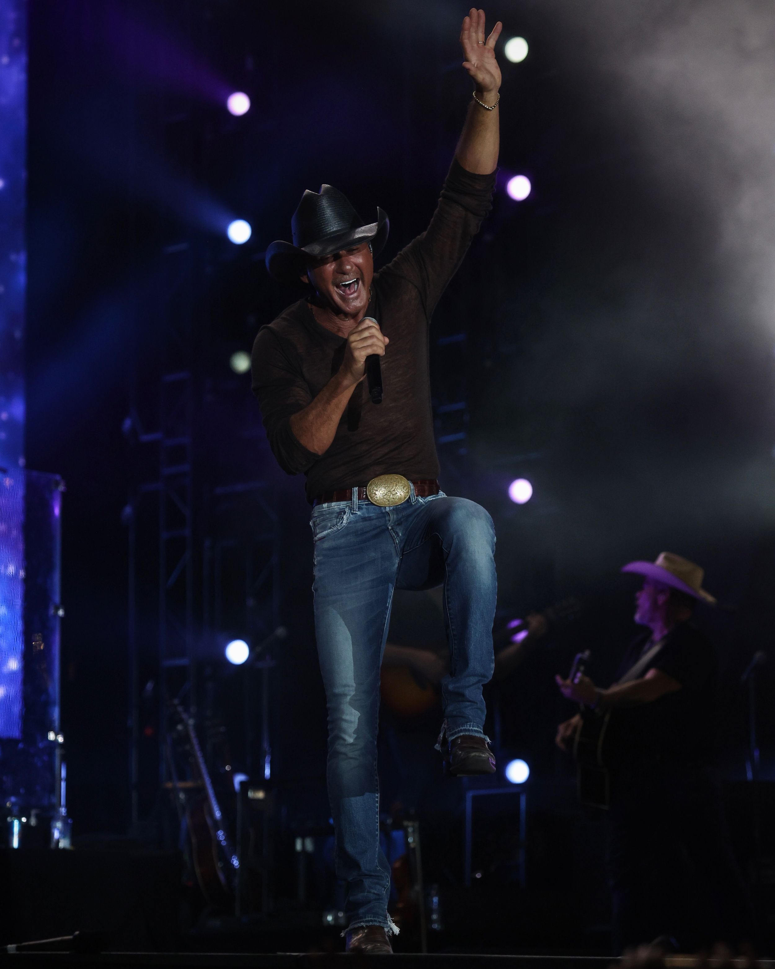 Country performer in cowboy hat and jeans singing energetically on dimly lit concert stage.