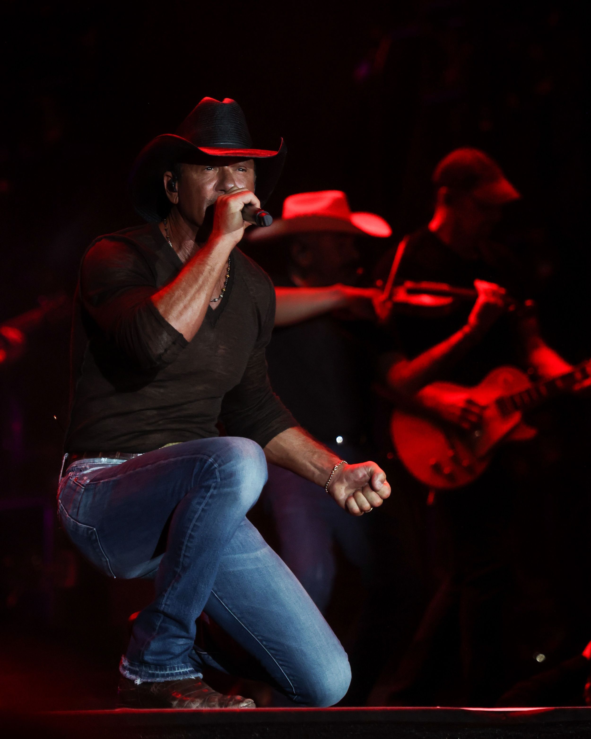 Country singer in black shirt and cowboy hat performs on stage with red lighting.