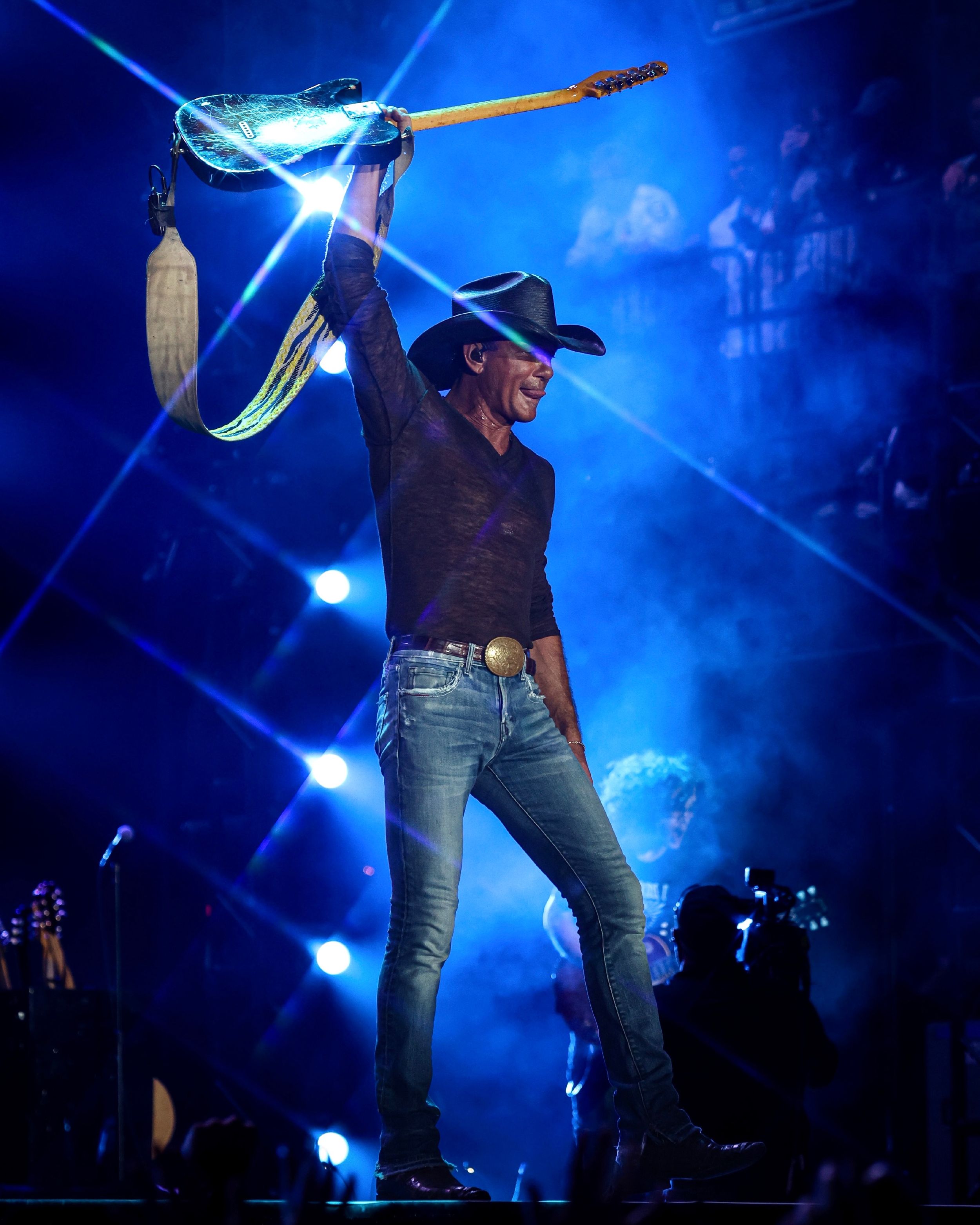 Country performer in cowboy hat holds guitar up while performing on stage with blue lighting.
