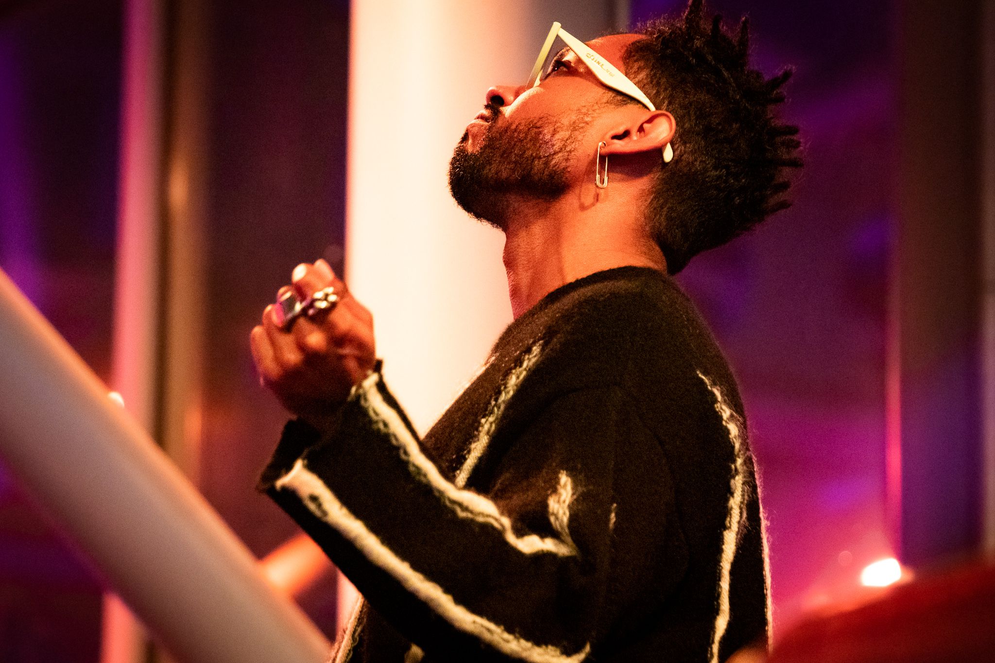 Performer passionately singing into microphone on stage with dramatic purple lighting in background.