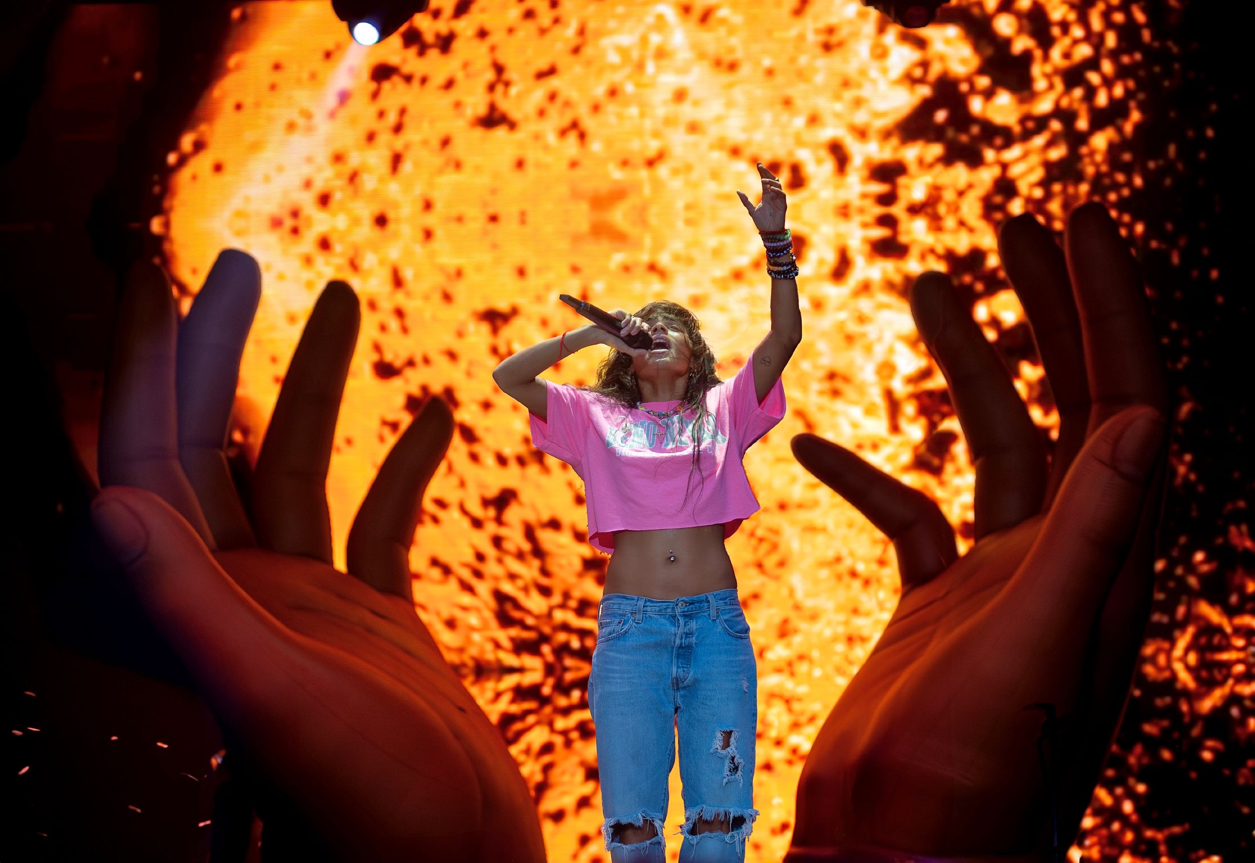 Stage performer with raised arms against explosive orange backdrop and giant hands.