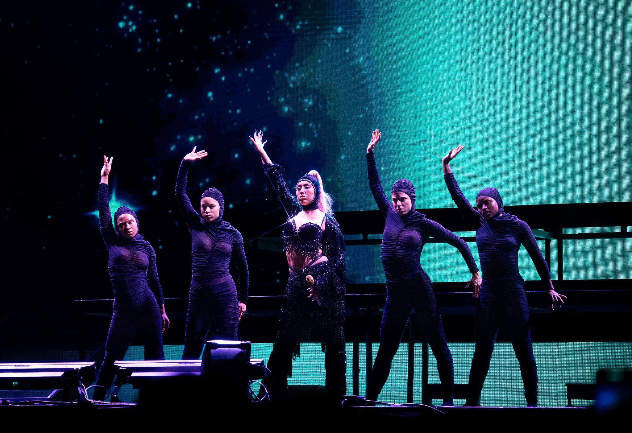 Performers in coordinated outfits dancing on stage with teal lighting effects.
