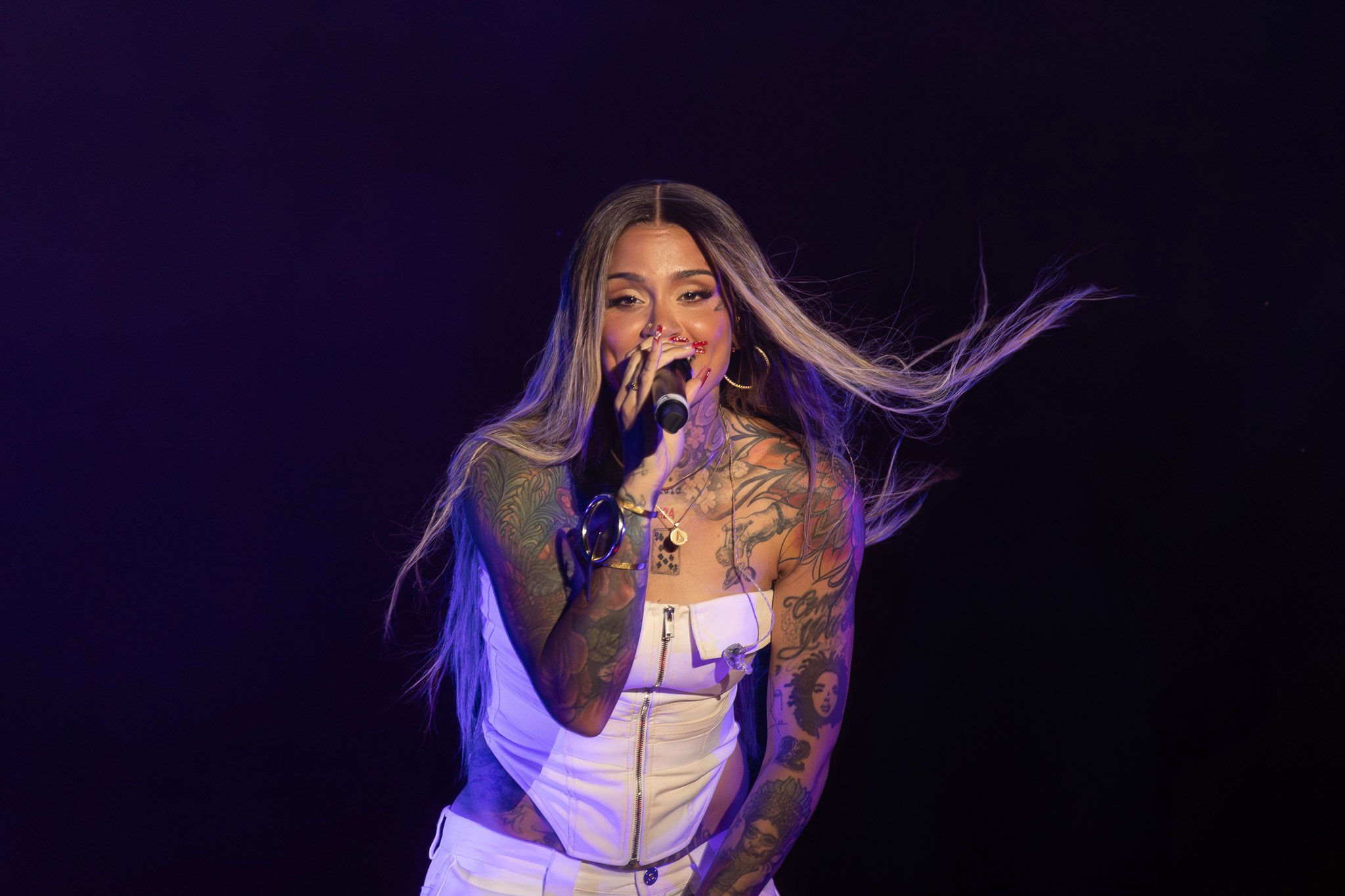 Concert performer in white outfit with flowing hair against purple backdrop.