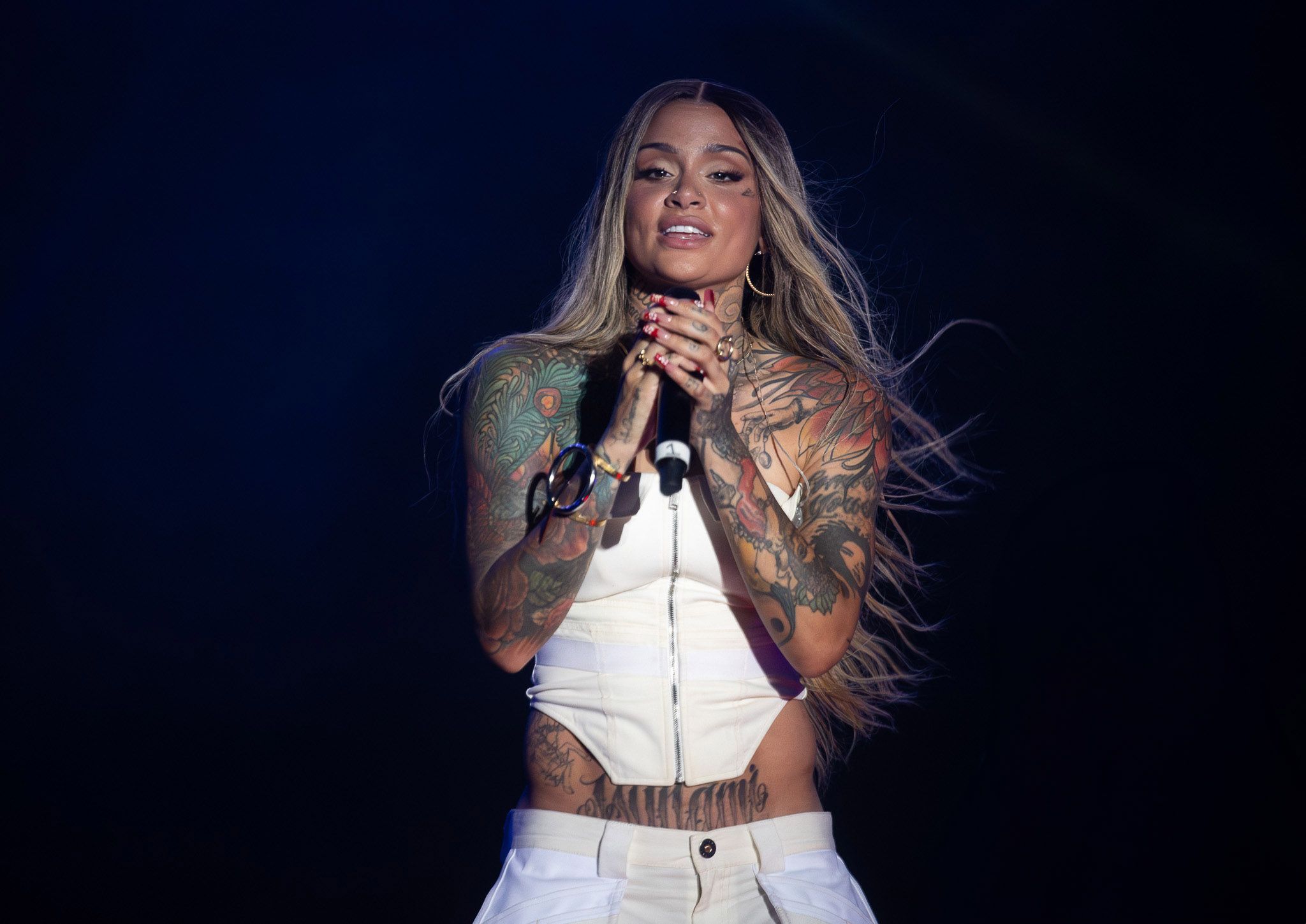 Stage performer in white outfit singing with flowing hair against dark background.