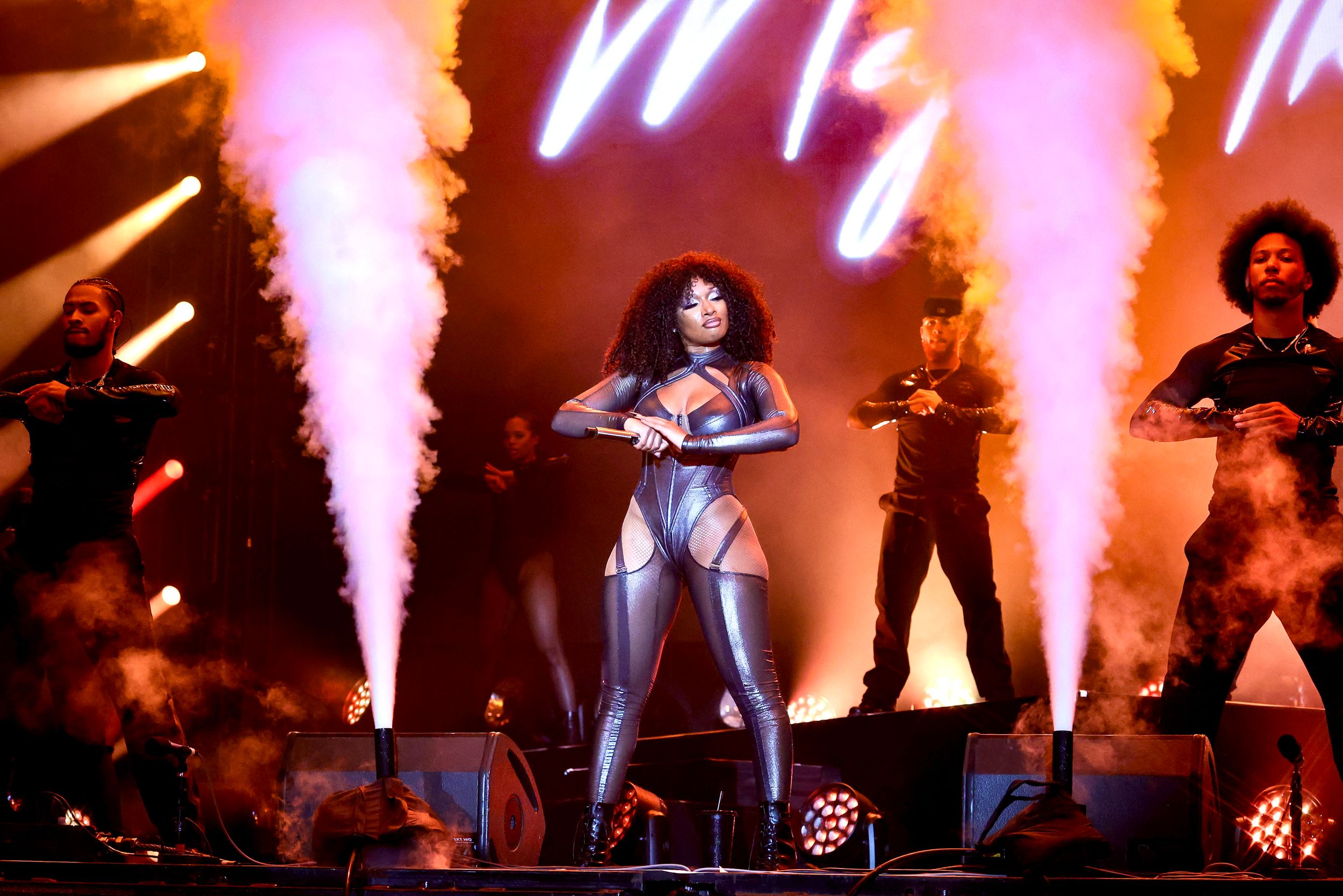 Concert performer in silver outfit with backup dancers and pyrotechnics on stage.