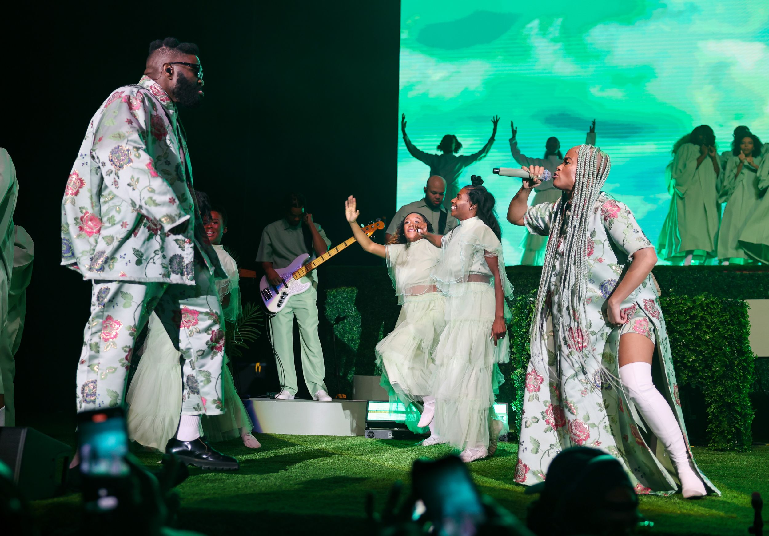 Performers in white and floral outfits dancing on stage with artificial grass and turquoise backdrop.