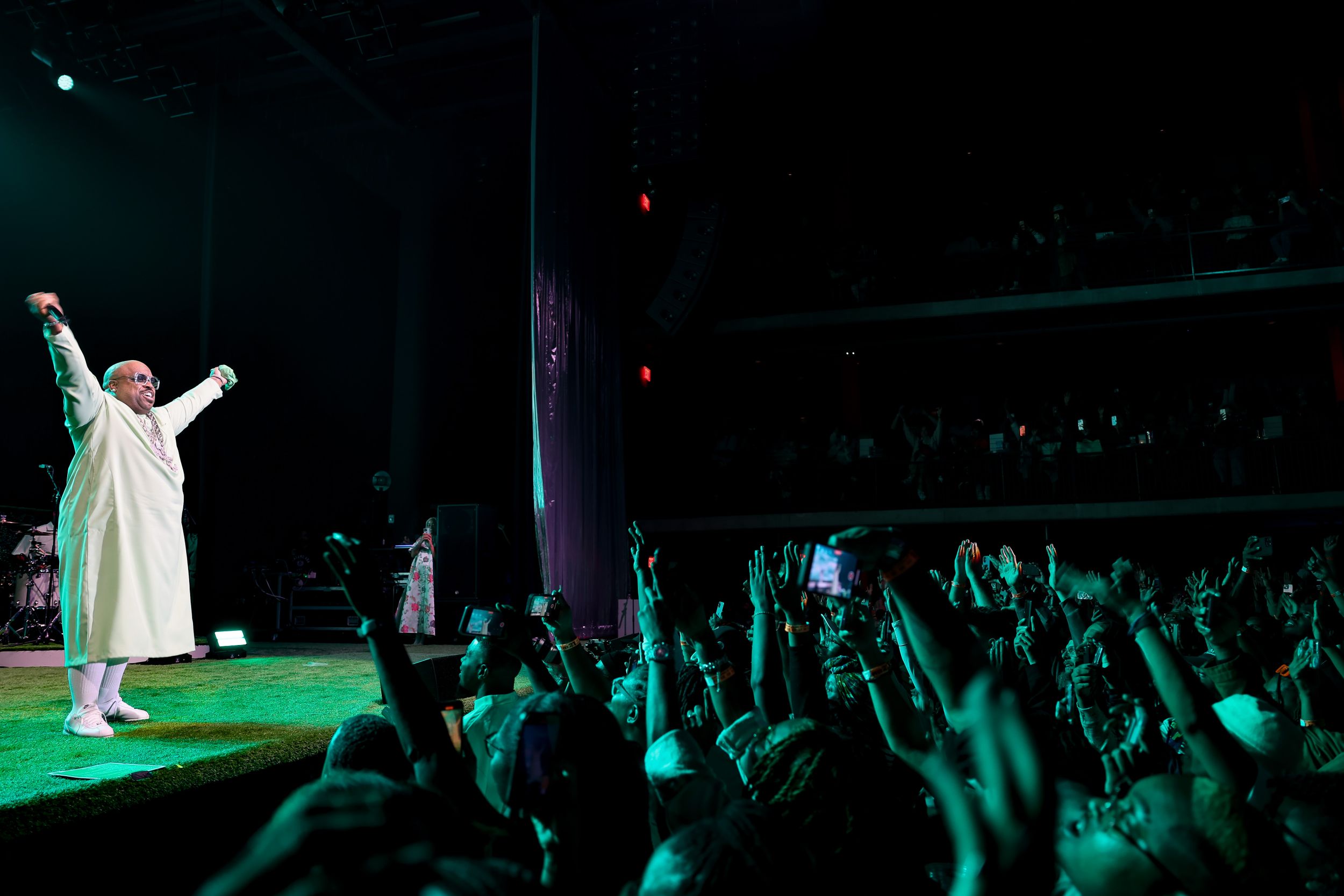 Concert performer in white robe on stage with green lighting and crowd raising hands in silhouette.