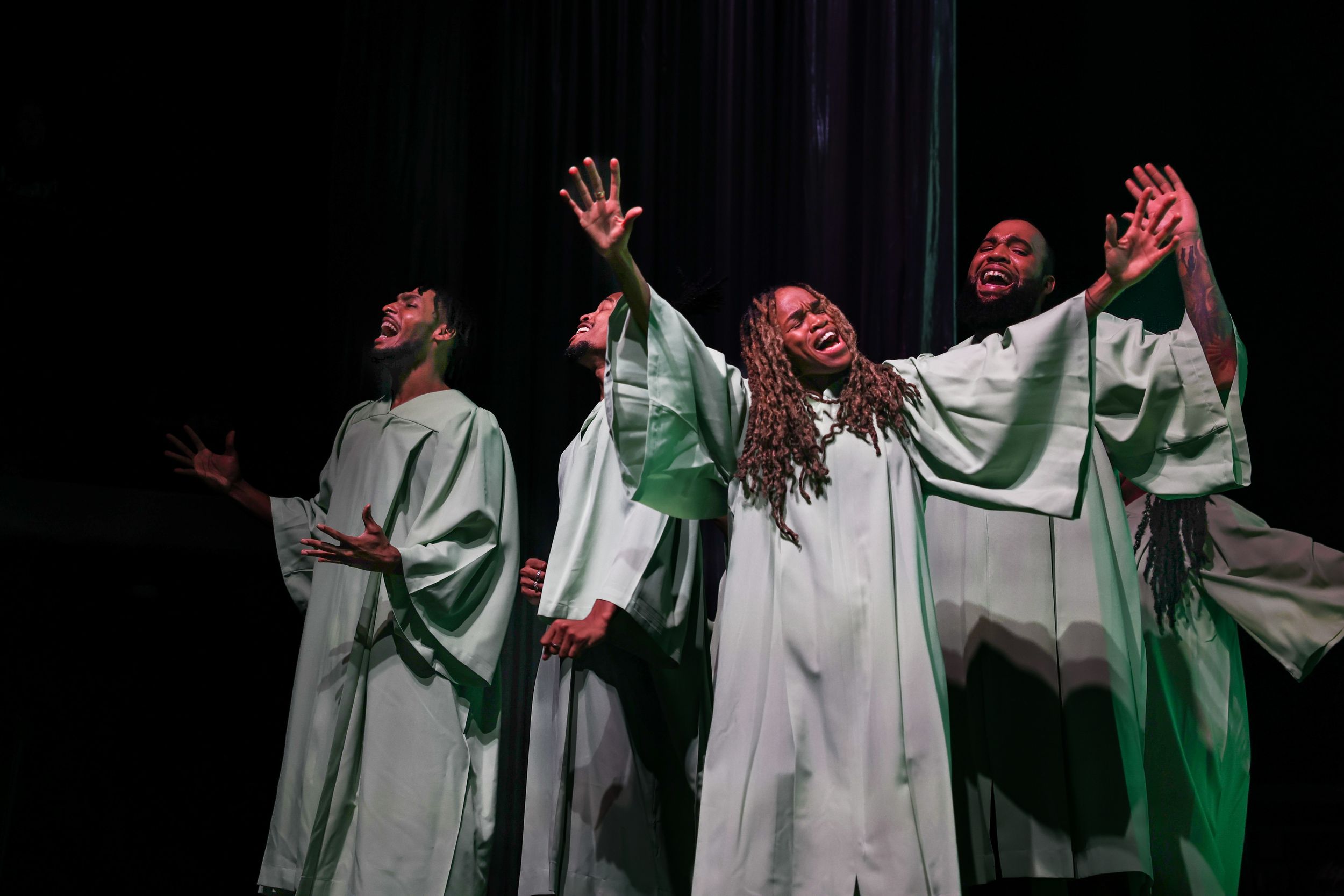 Gospel choir members in white robes perform with raised hands under theatrical lighting.