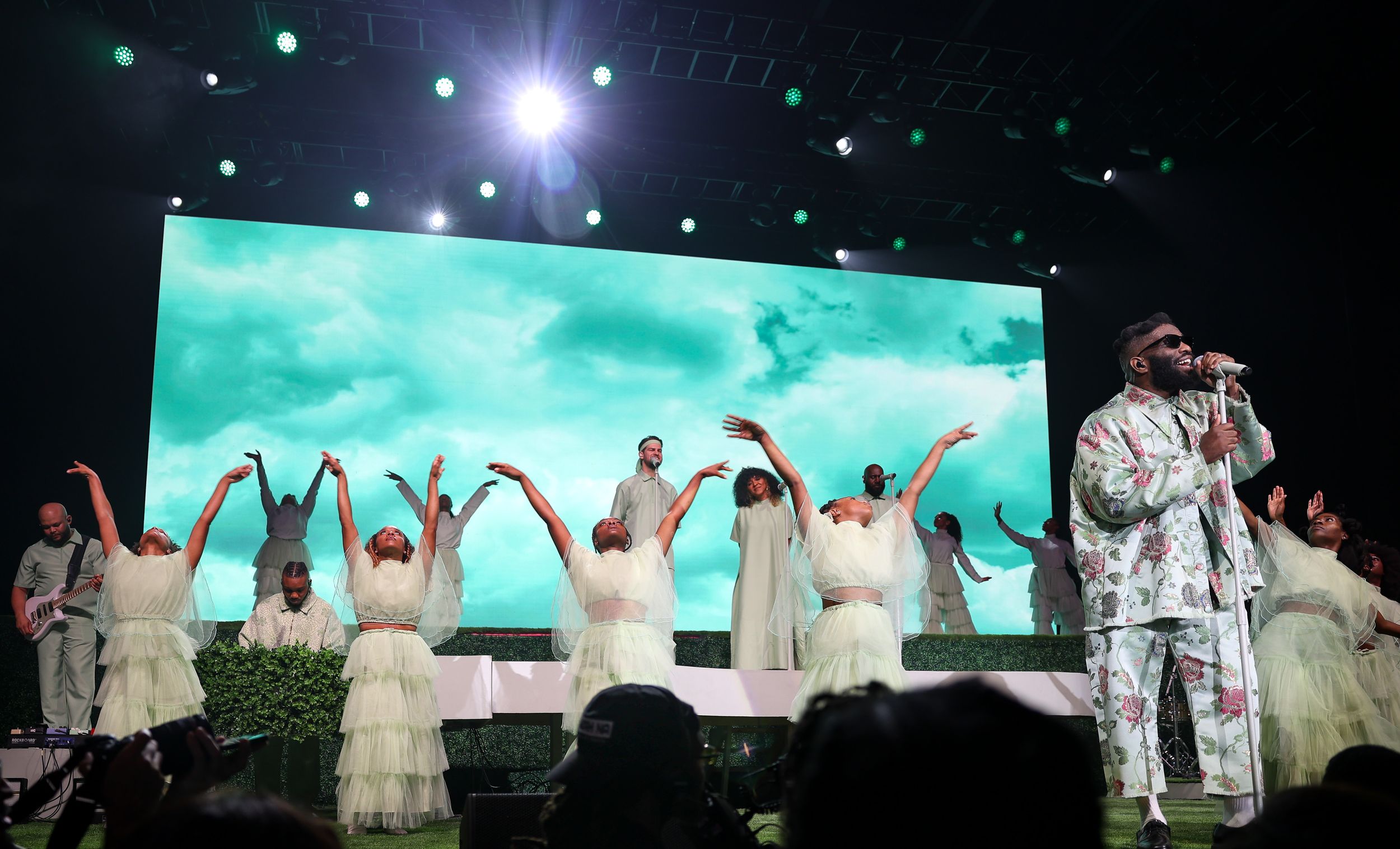 Performance on stage with dancers in white dresses against turquoise backdrop raising arms in celebration.