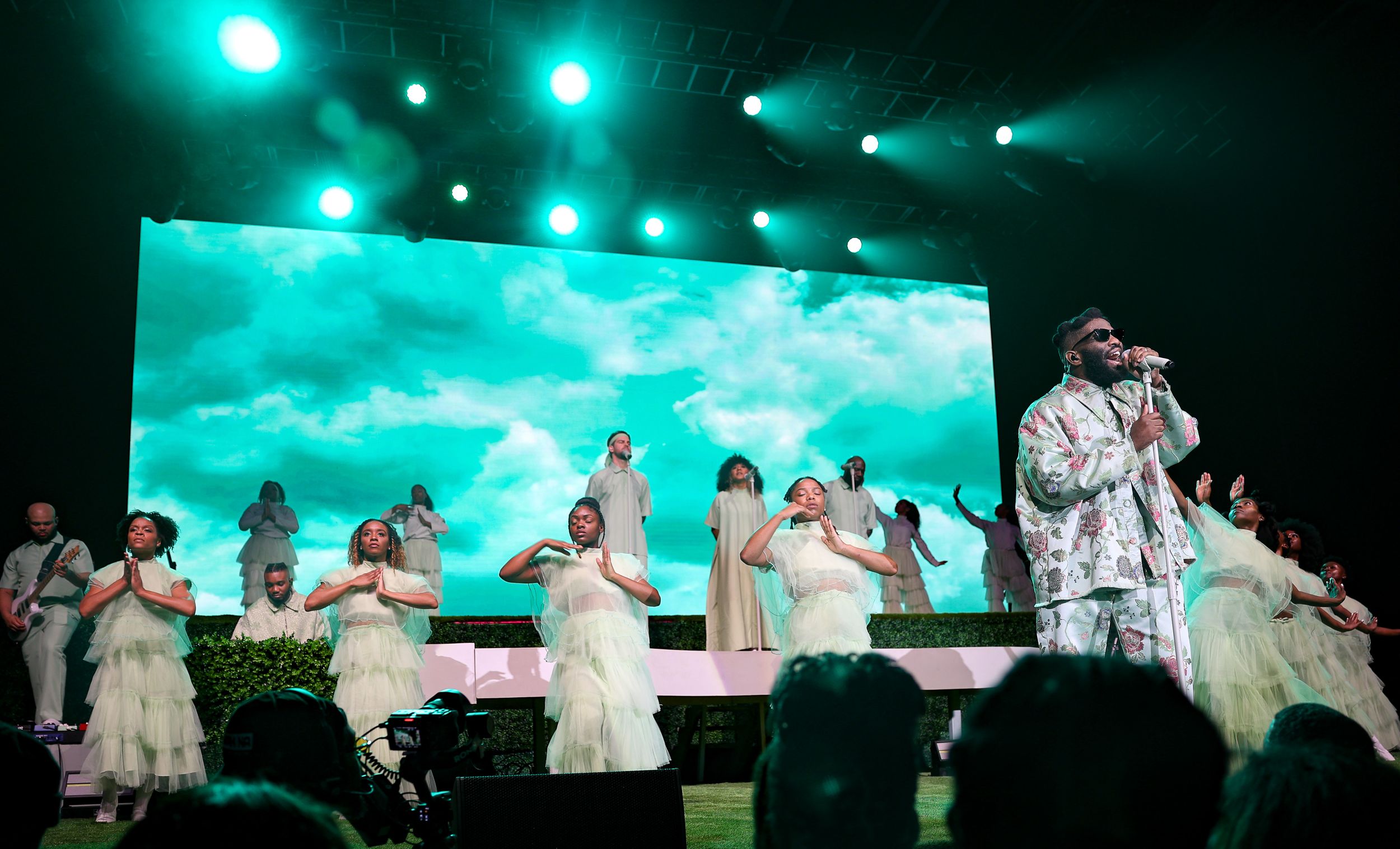 Concert performers in white outfits on stage with turquoise backdrop and dramatic lighting during live performance.