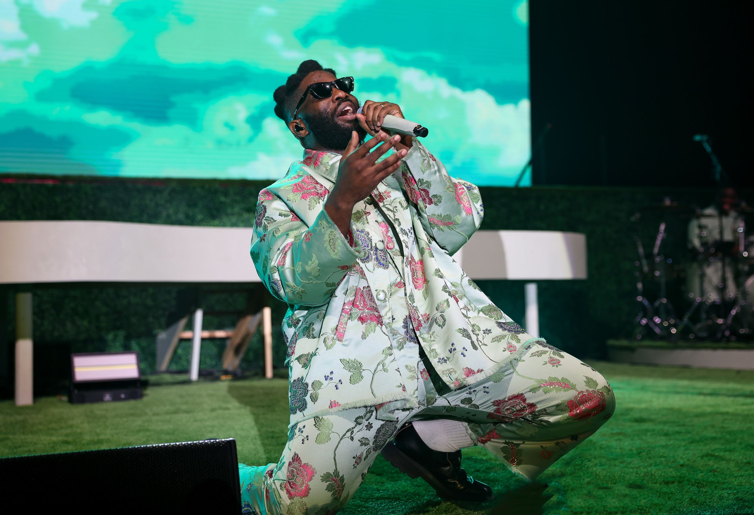 Performer in floral patterned outfit sings on stage with vibrant turquoise backdrop.