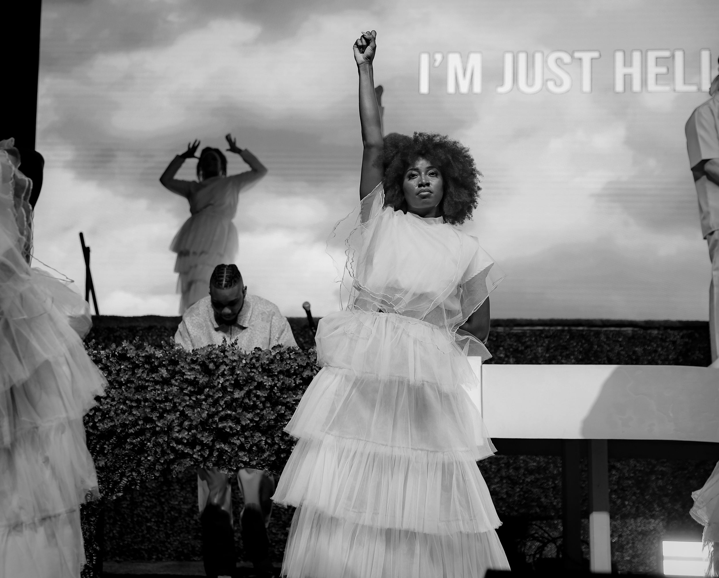 Black and white photo of performers in white dresses with text overlay reading I'M JUST HELP.