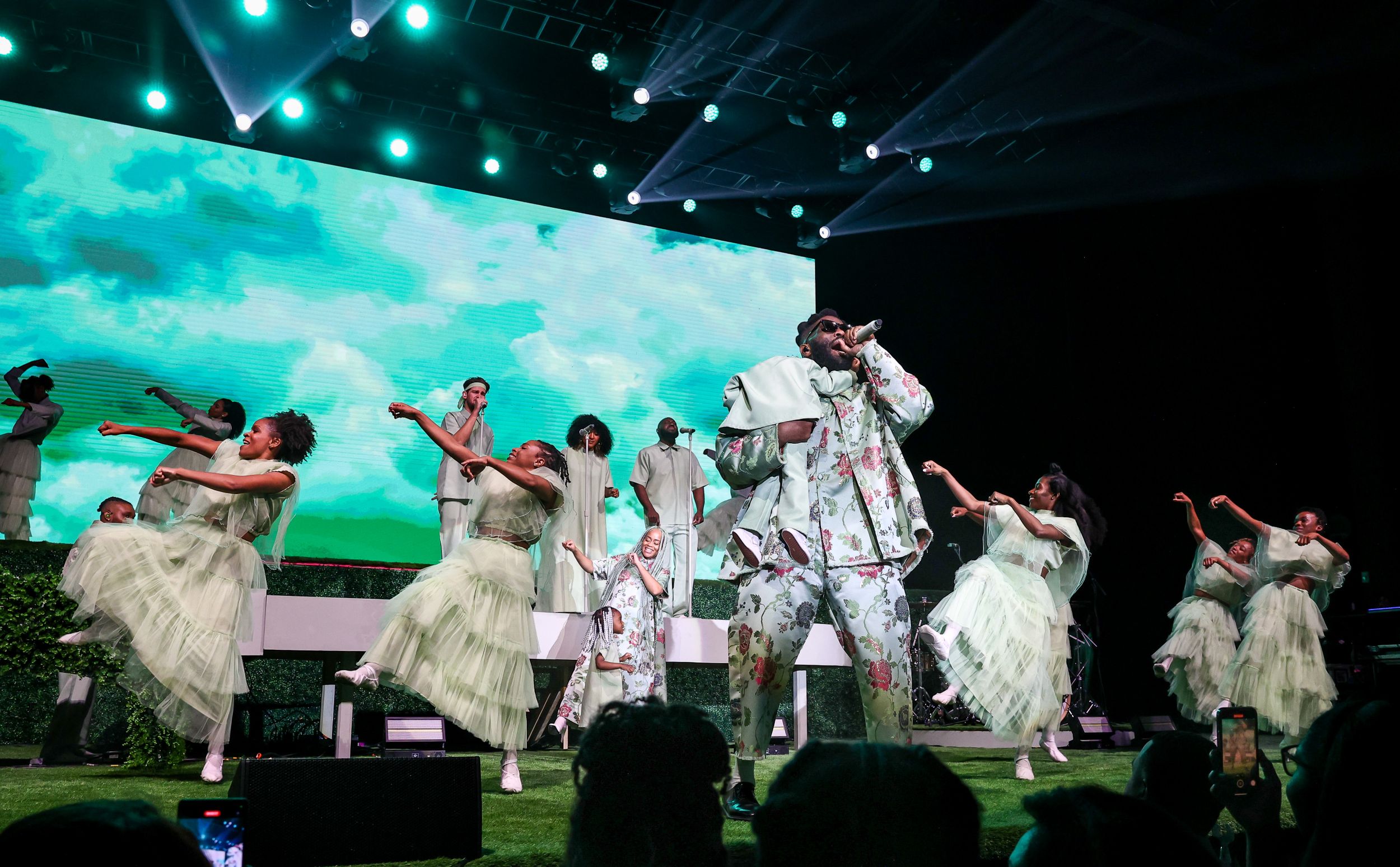 Performers in flowing white dresses dance on stage with dramatic turquoise backdrop.