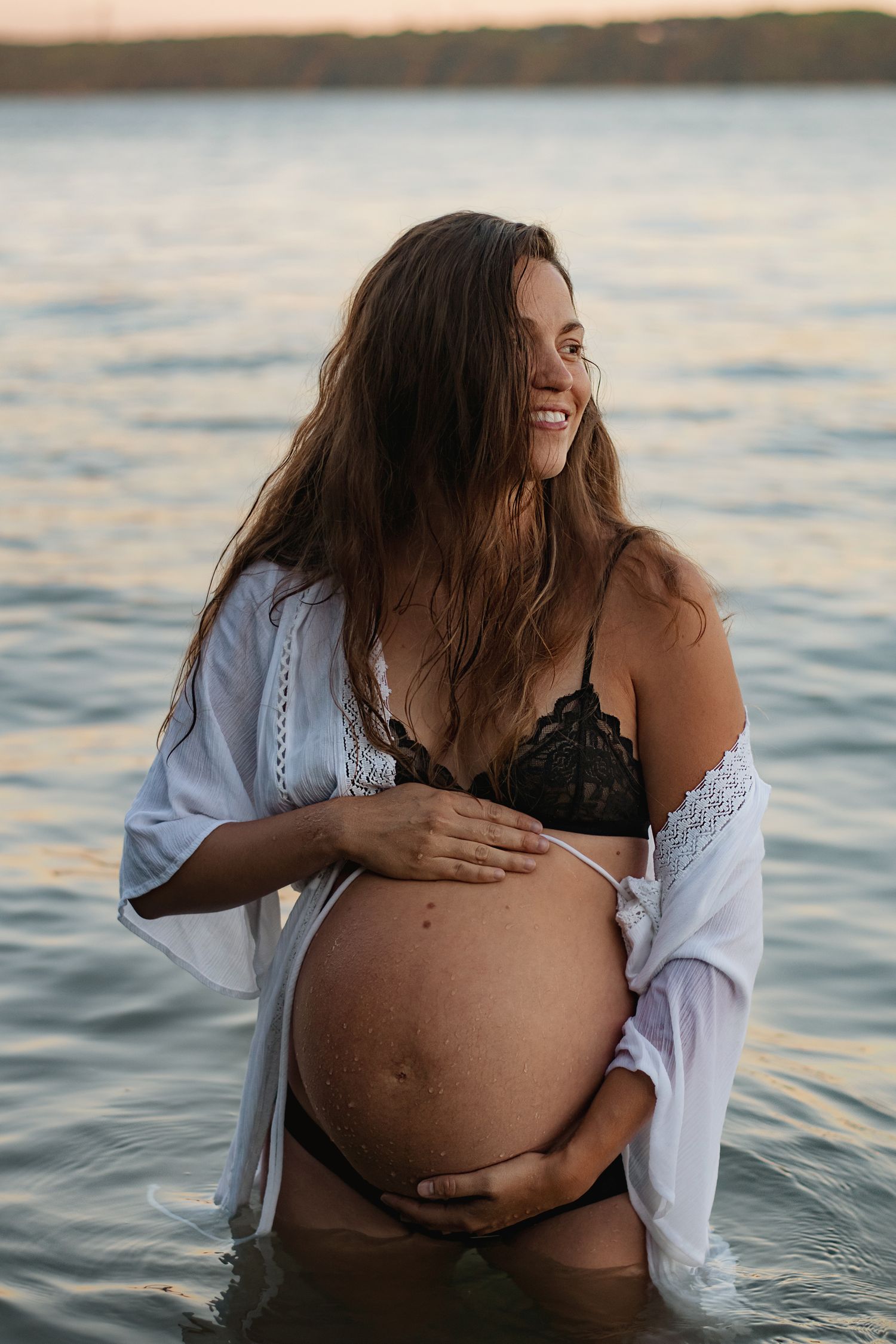 Maternity photo of woman in white shirt and black swimsuit standing in water at sunset.