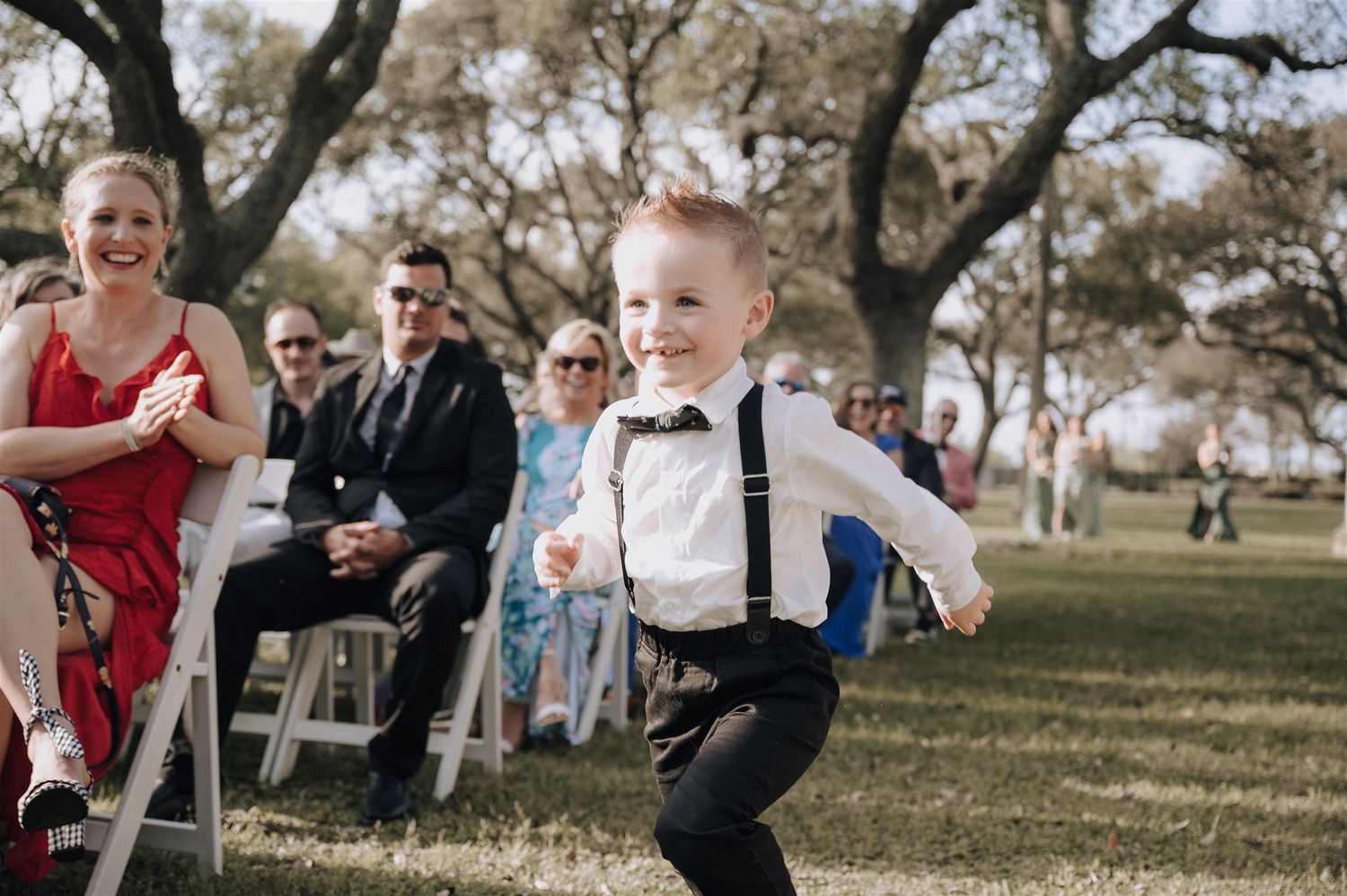 A young child in suspenders runs joyfully down the aisle at an outdoor wedding.