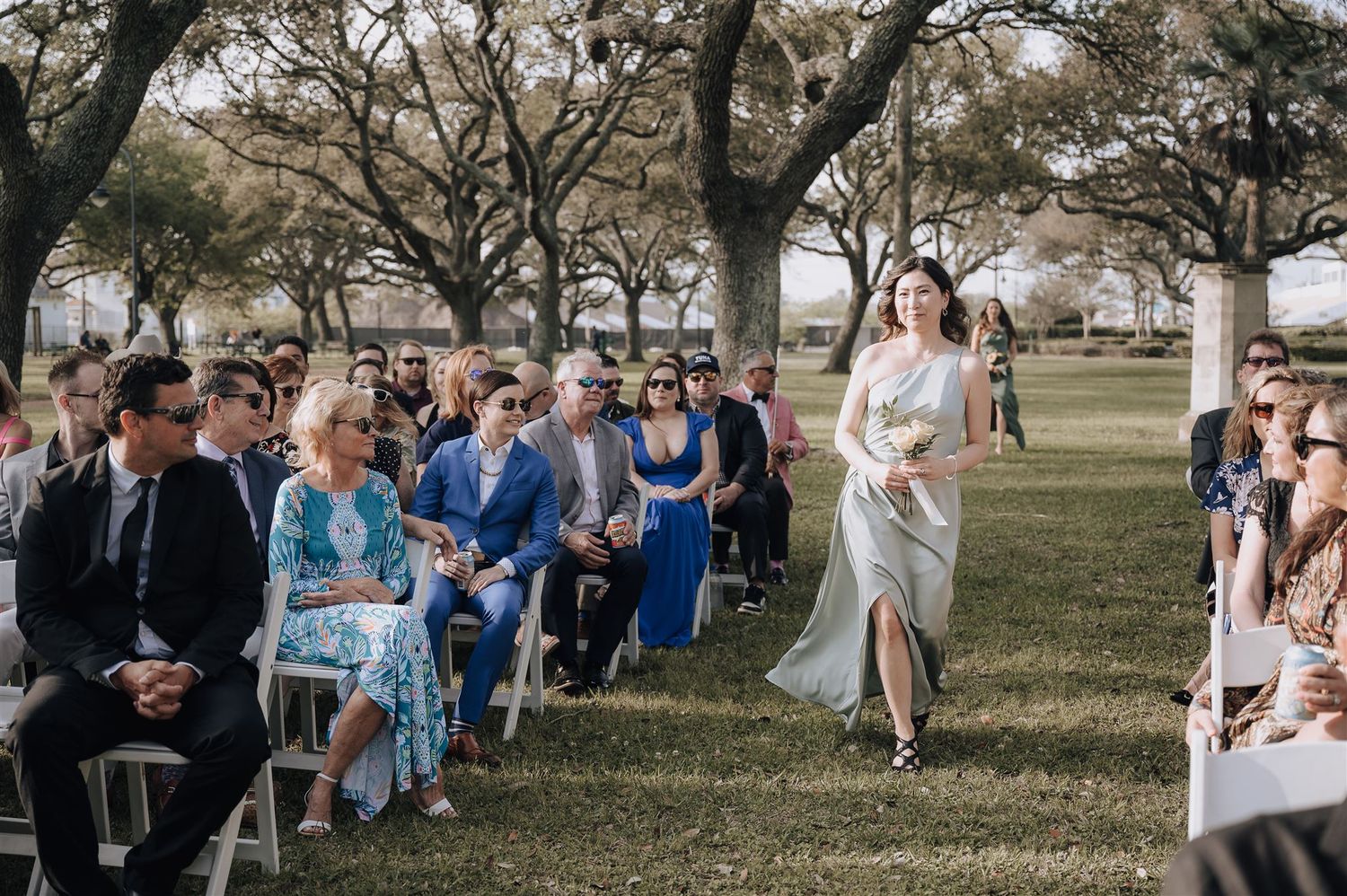 A wedding attendant proceeds down the aisle at an outdoor ceremony with guests seated on white chairs.