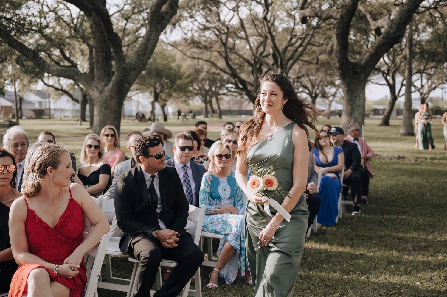 A bridesmaid carries a bouquet while walking past seated guests at an outdoor wedding.