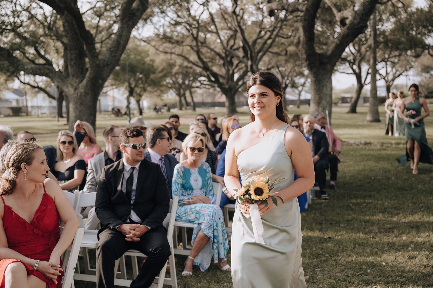 A bridesmaid in a light-colored dress walks down a grassy aisle at an outdoor ceremony.