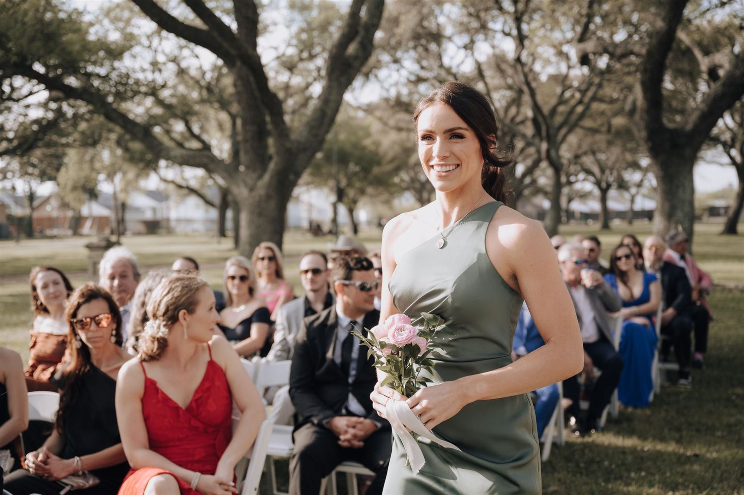 A bridesmaid in a sage green dress walks down the aisle at an outdoor wedding ceremony.