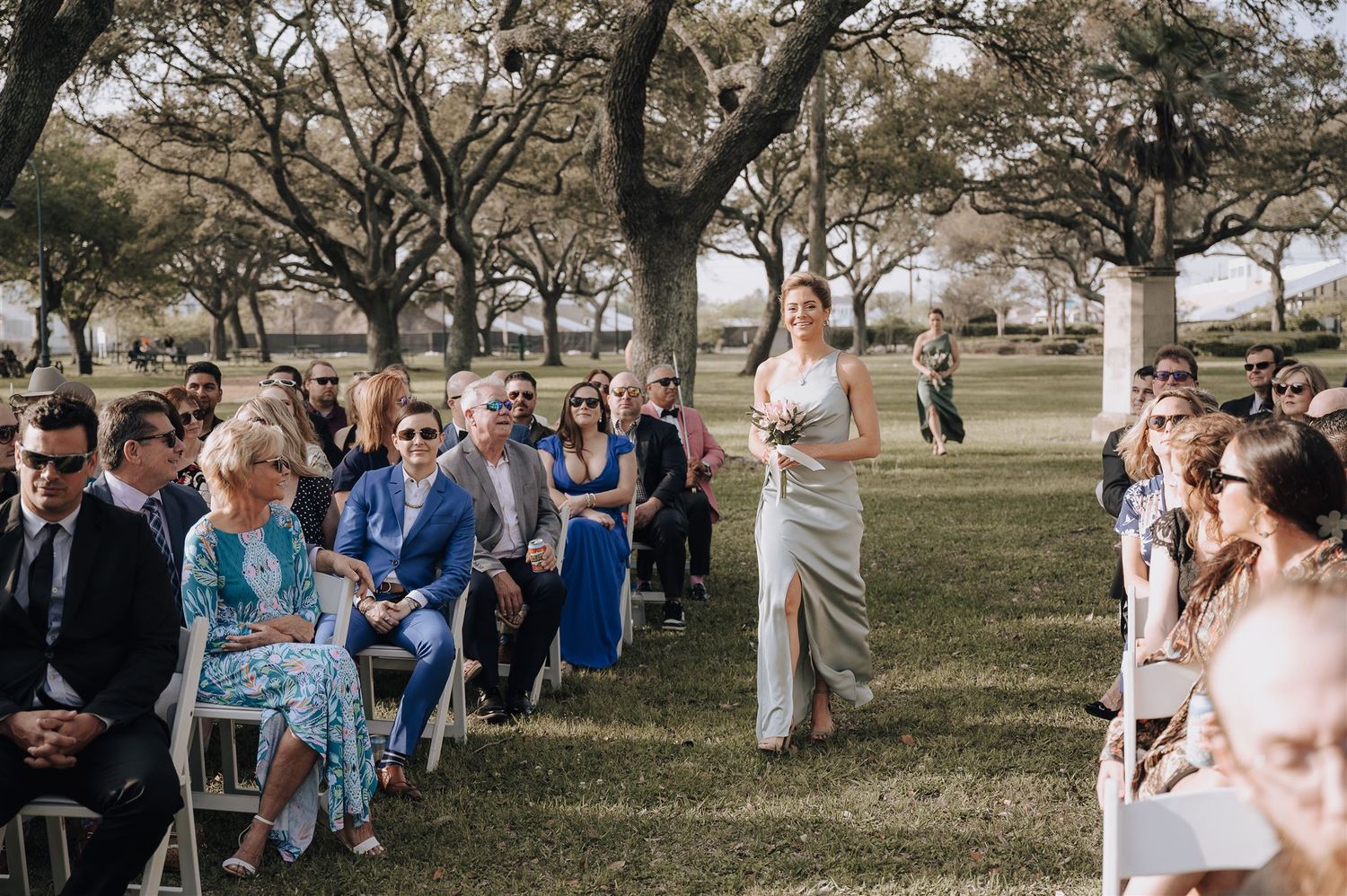 Wedding guests seated on white chairs at an outdoor ceremony beneath sprawling oak trees.