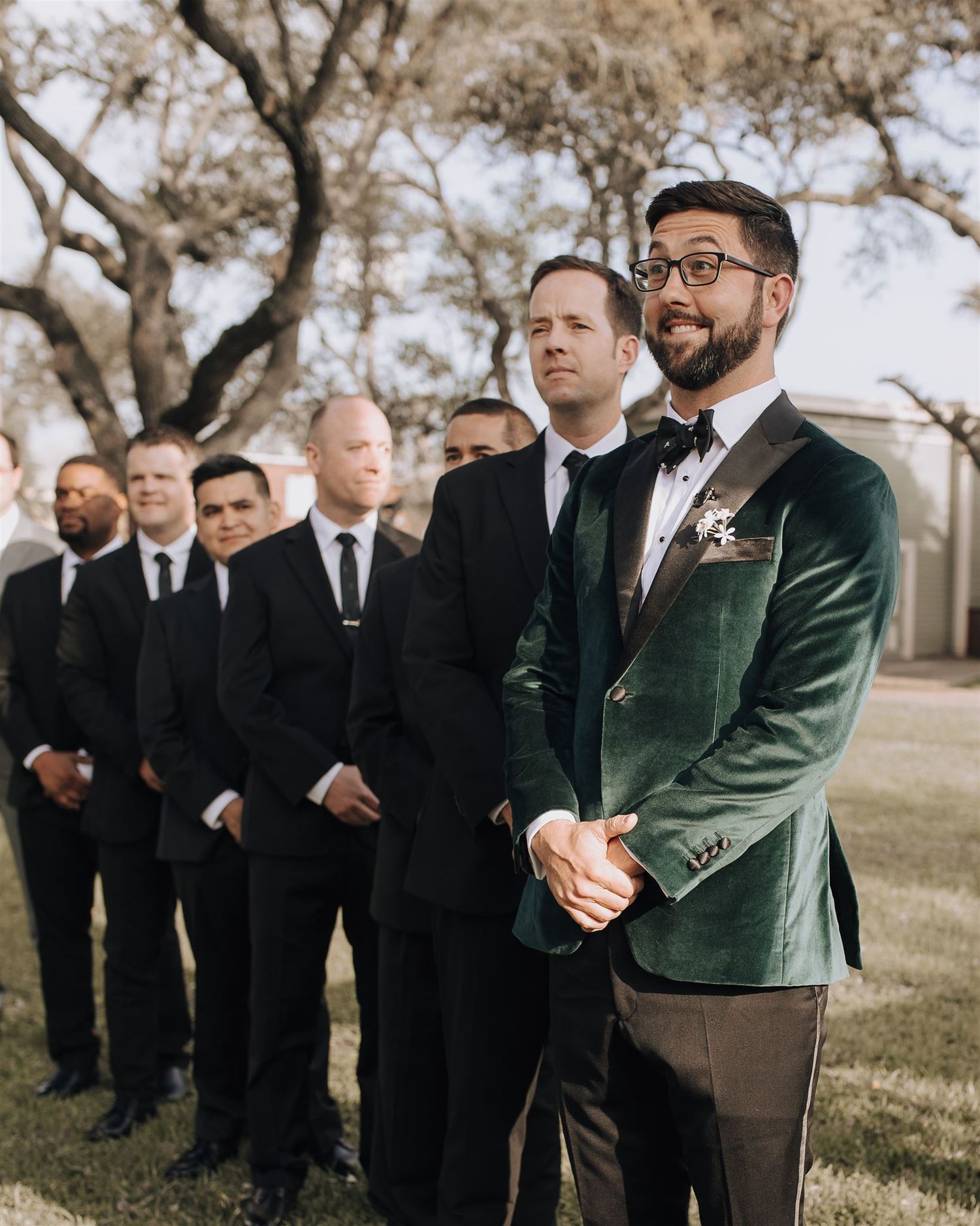 Groomsmen in dark suits stand in line at an outdoor wedding with one wearing a green velvet jacket.