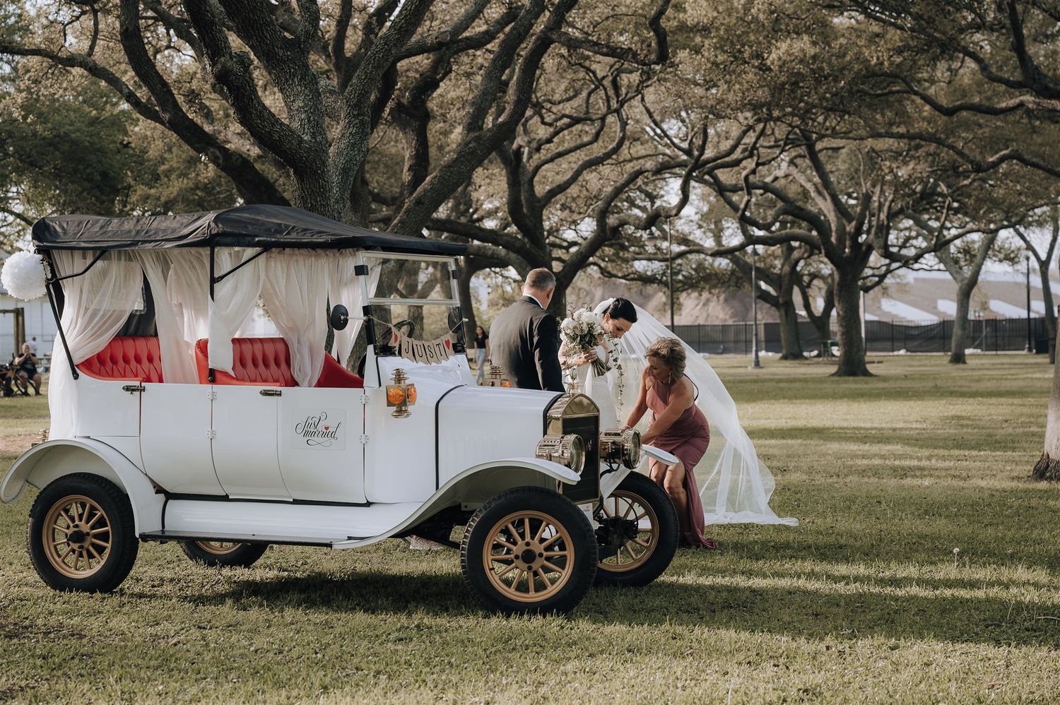 A vintage white car serves as a backdrop for a wedding ceremony under oak trees.