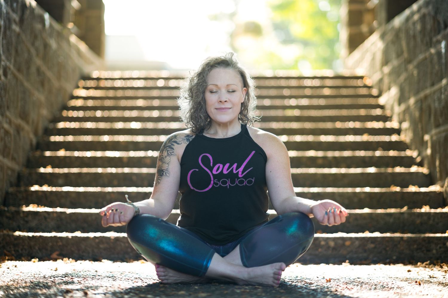 Fitness instructor sitting criss cross on concrete stairway landing with hands rested upwards on knees eyes closed smile