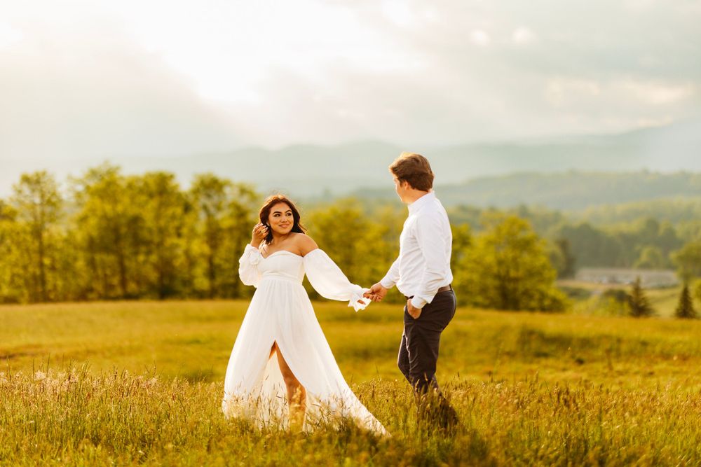 Bride in flowing white dress holds grooms hand walking through sunlit green mountain meadow.