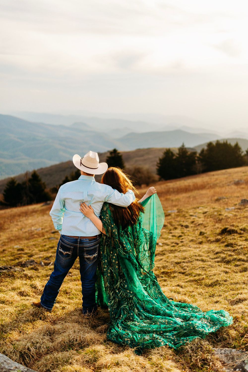 Couple embracing from behind on mountain hillside, woman in green dress, man in white cowboy hat, scenic valley view.
