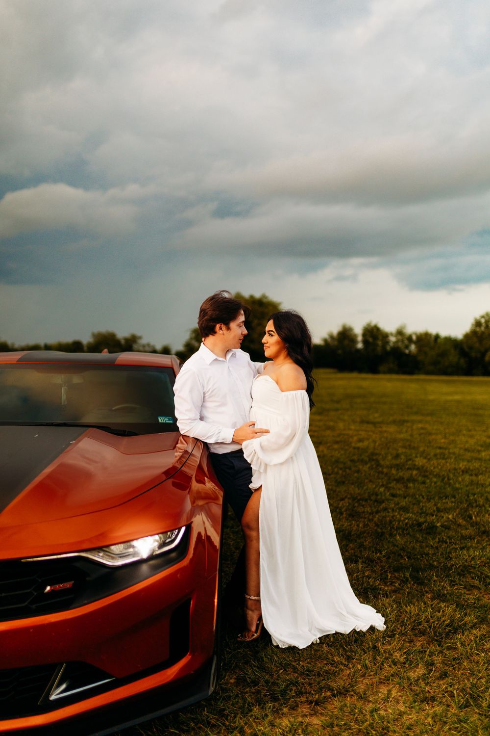 Couple embracing beside a red Chevrolet Camaro in open field under dramatic cloudy sky at sunset.