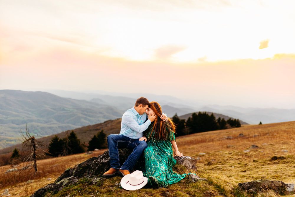 Couple sharing intimate moment on hillside rock at sunset, woman in emerald dress, white hat on ground nearby.