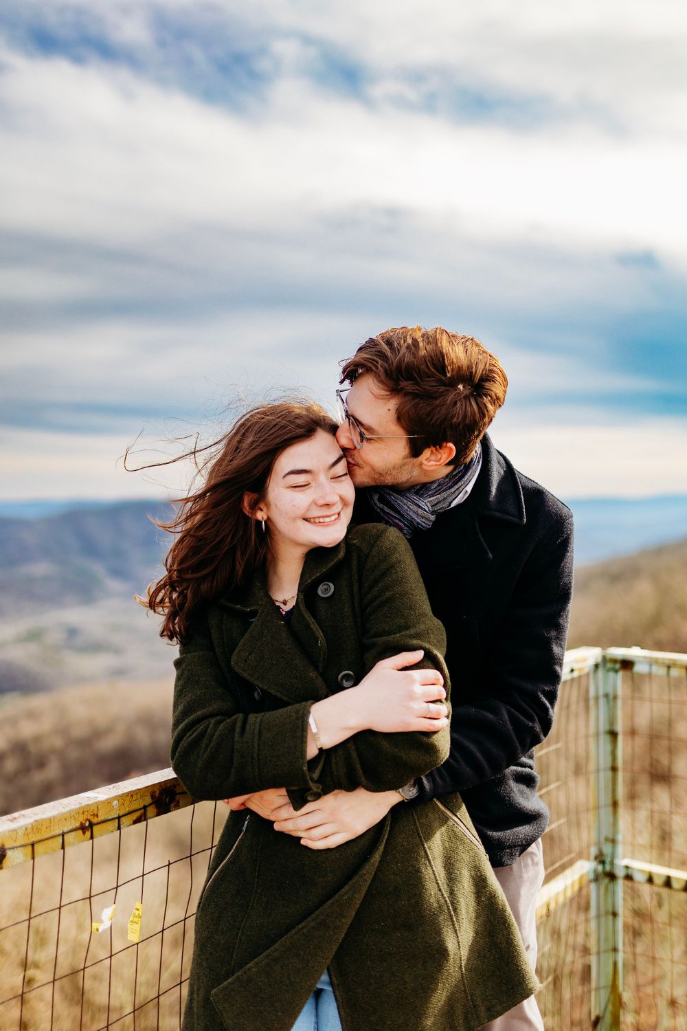Man kissing smiling woman on cheek while hugging her on a windy mountain overlook deck.