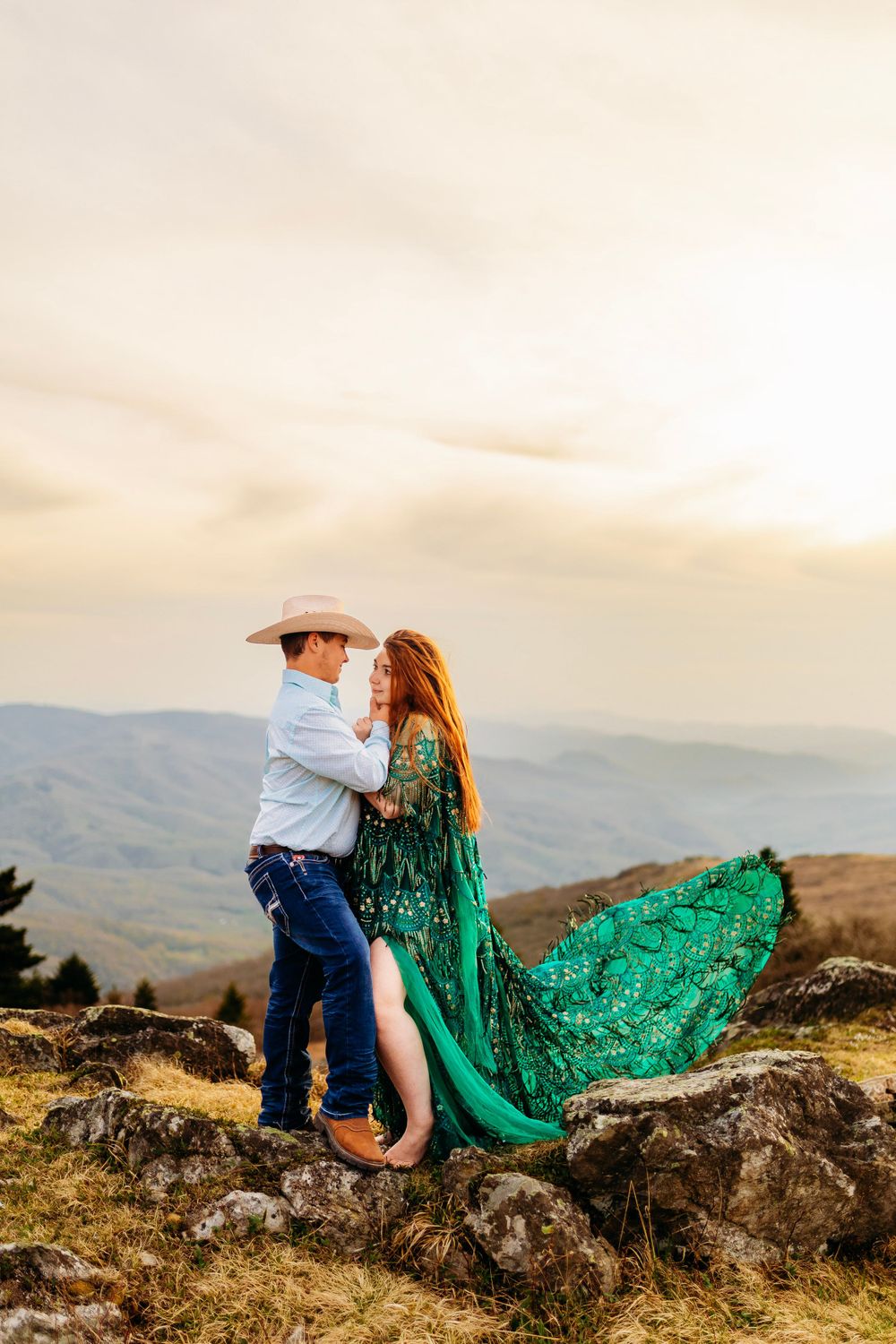 Couple embracing on rocky mountain overlook at sunset, woman in flowing green dress, man in white cowboy hat.
