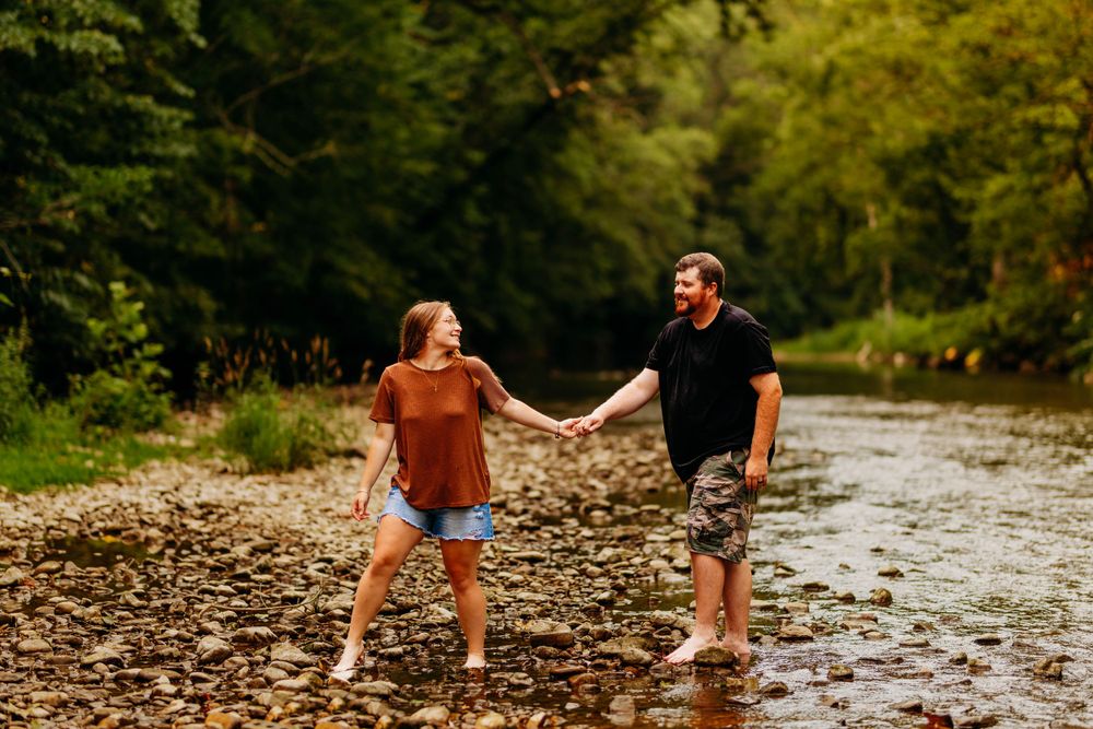 Couple holding hands and smiling at each other while wading in a rocky creek surrounded by lush green trees.