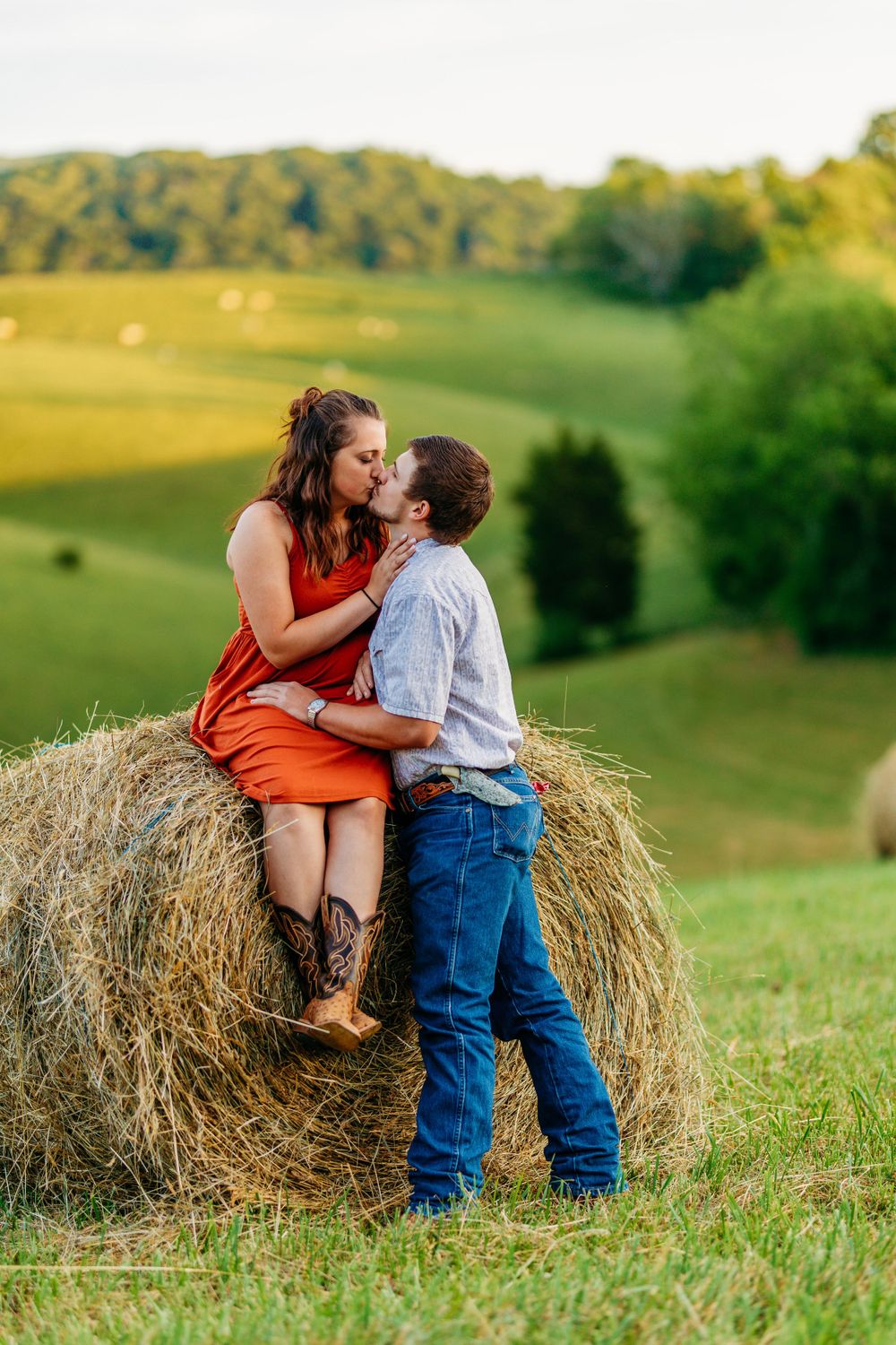 Couple sharing a romantic kiss on a hay bale in a lush green countryside at golden hour.
