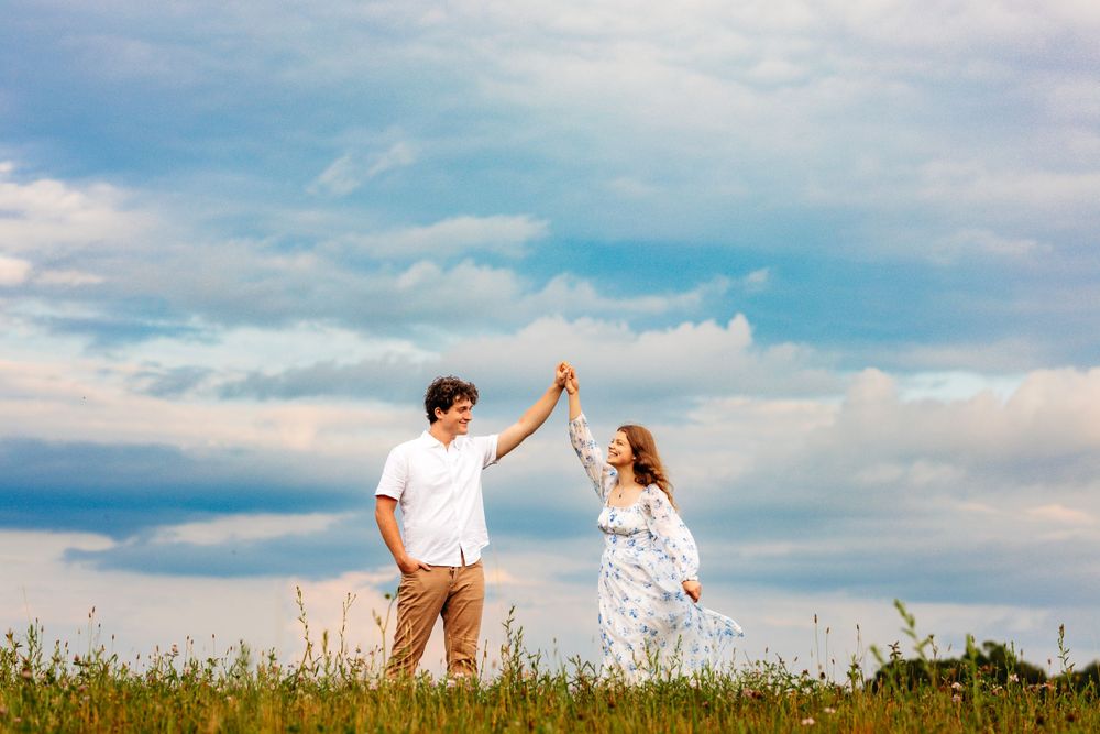 Couple dancing together in sunny green meadow field, man spinning woman in white dress under blue cloudy sky.