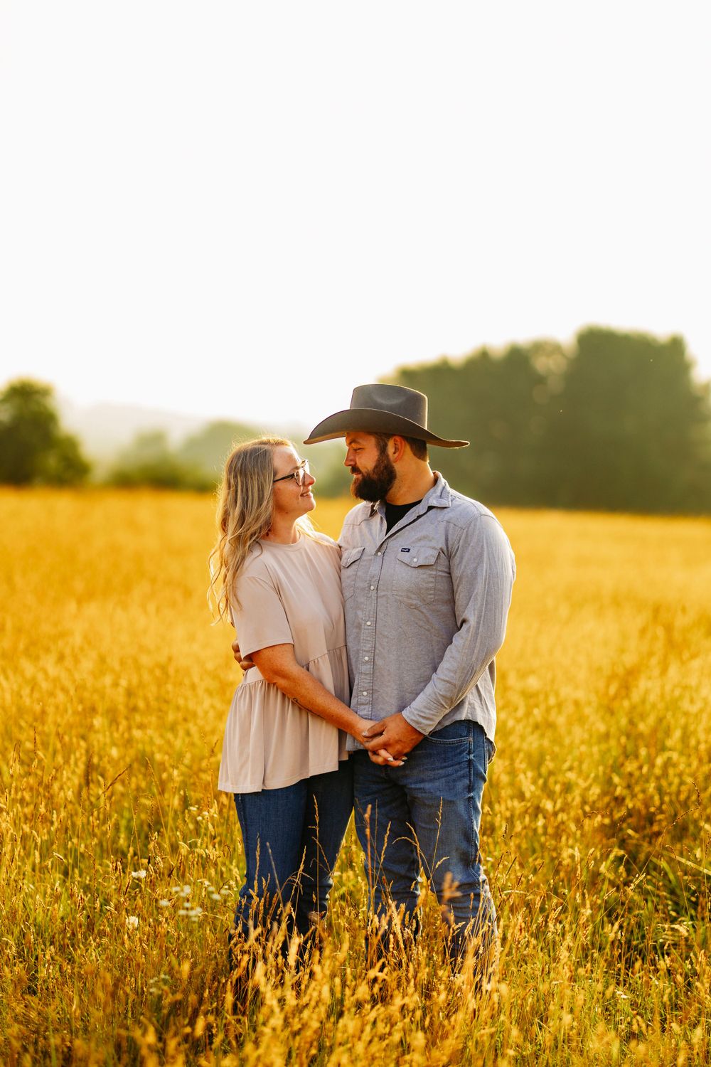 Couple holding hands gazing at each other in a sunlit golden grain field.