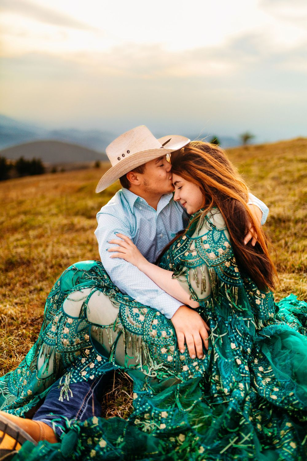 Couple sitting in golden meadow at sunset, man in cowboy hat embracing woman in green boho dress.