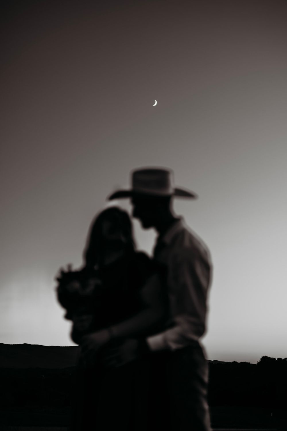 Romantic couple silhouette embracing under a crescent moon at dusk in a moody black and white engagement photo.