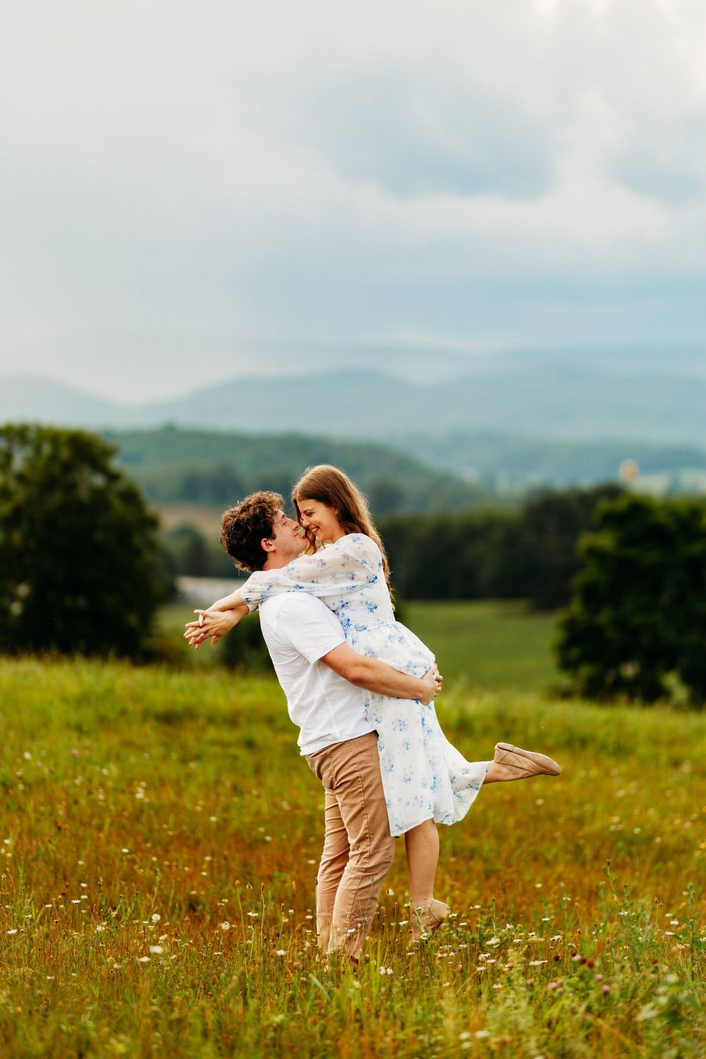 Romantic couple embracing in lush green meadow with scenic mountain landscape and dramatic cloudy sky backdrop.