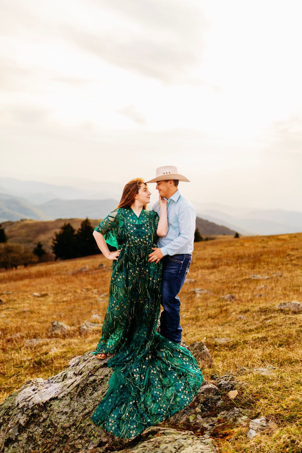 Couple standing on rocky mountain overlook, woman in green dress and man in cowboy hat facing each other.